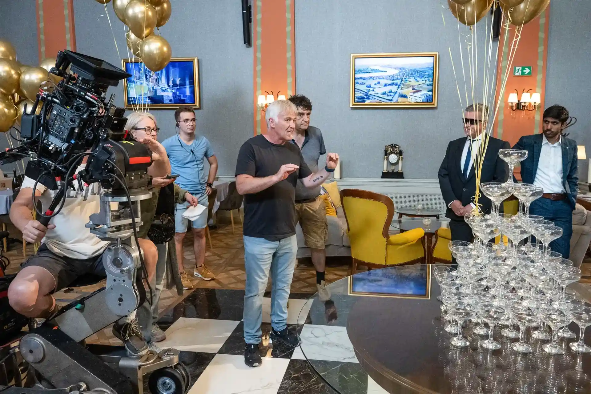 Film crew setting up a scene with a camera and a pyramid of champagne glasses in an elegant room with people preparing and discussing.