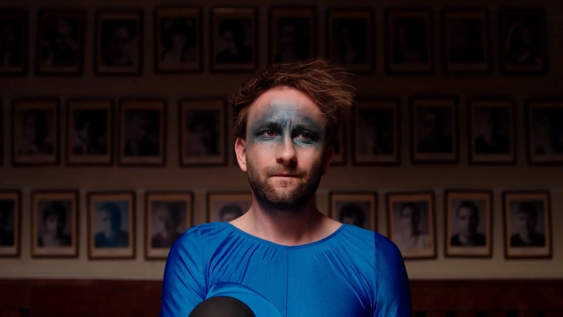 Man with dark blue face paint around eyes wearing a bright blue shirt, sitting in front of a wall with framed black-and-white portraits.