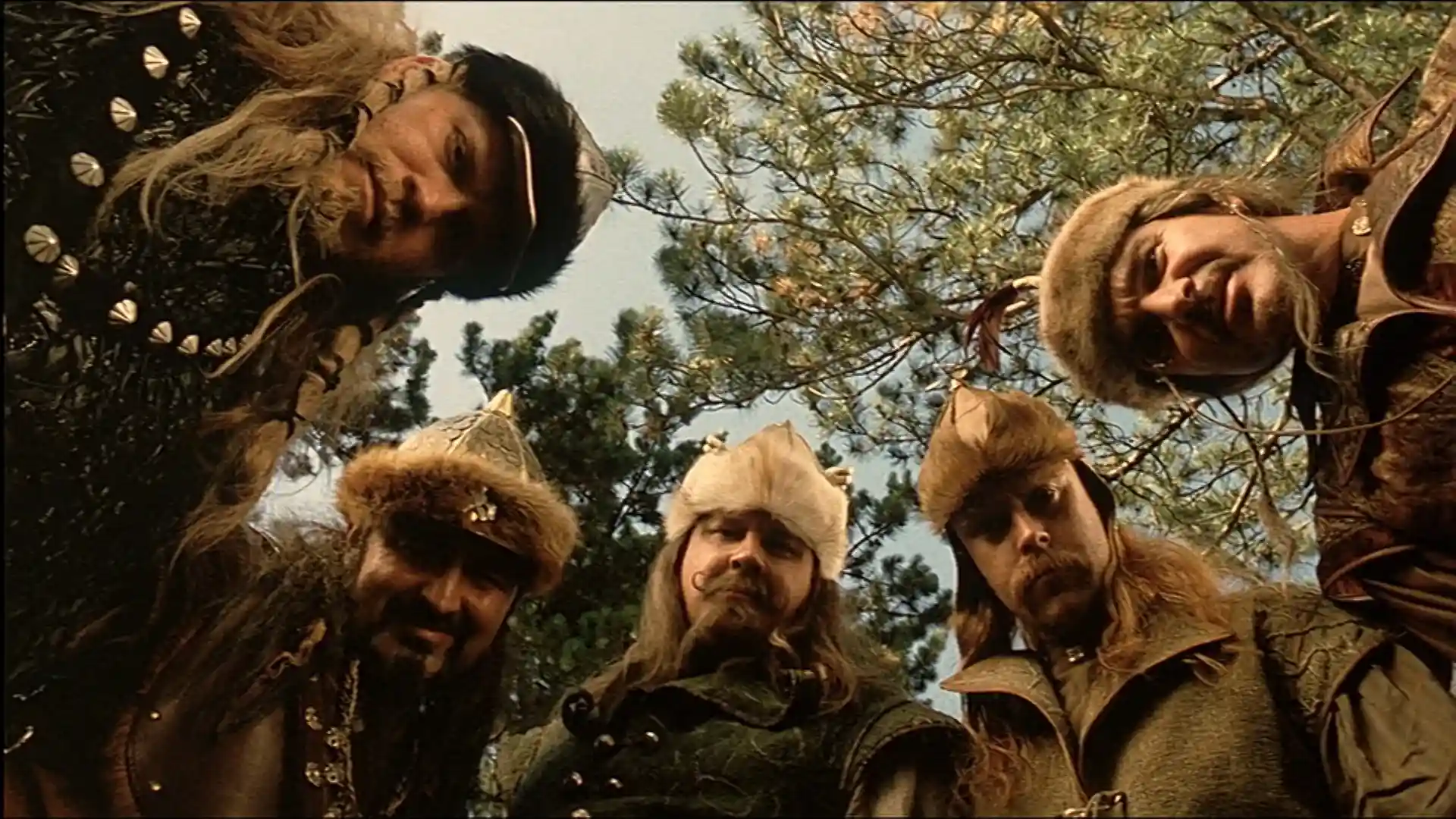 Five men dressed in medieval tribal warrior costumes and fur hats looking down at the camera with trees and sky in the background.