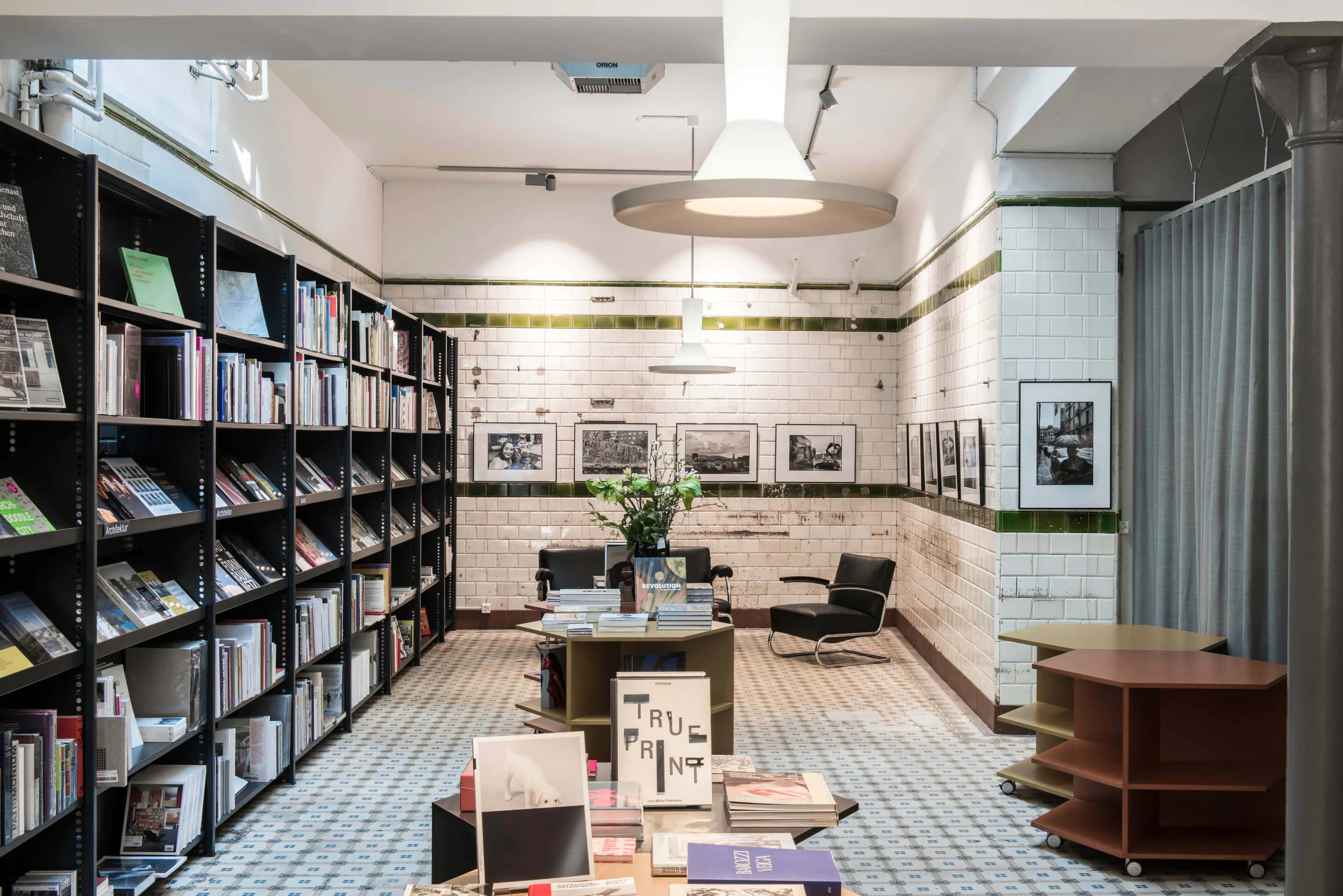 Innenraum einer Buchhandlung mit Regalen voller Bücher, Tischen mit gestapelten Büchern und schwarzen Sesseln vor einer weiß gekachelten Wand mit grünen Akzenten und schwarz-weißen Fotos.