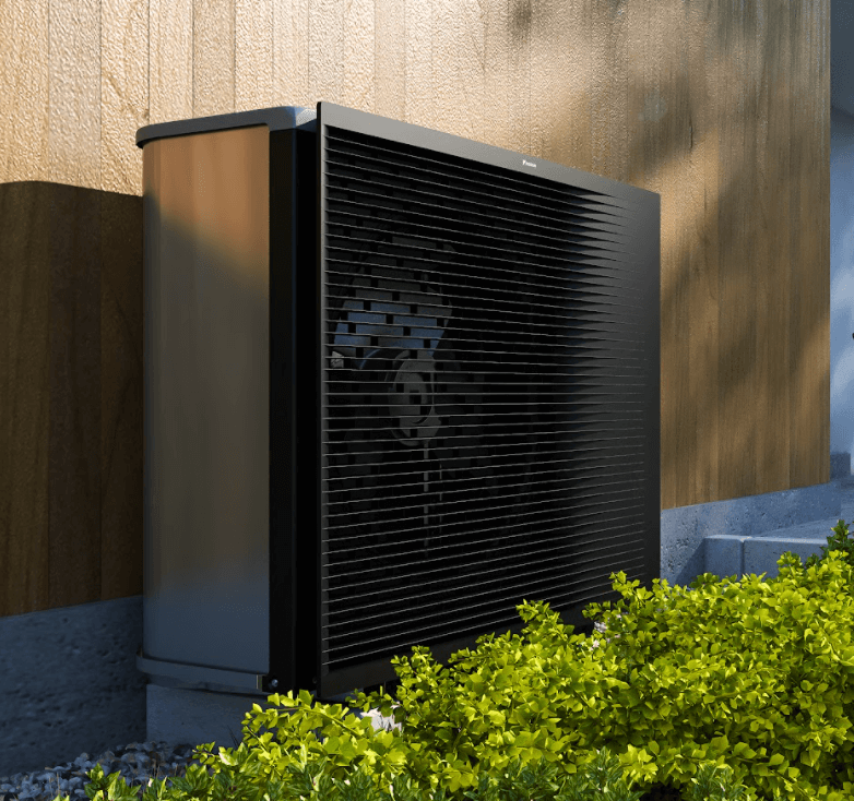 Unité extérieure d'une pompe à chaleur noire installée contre un mur en bois, avec des buissons verts au premier plan.