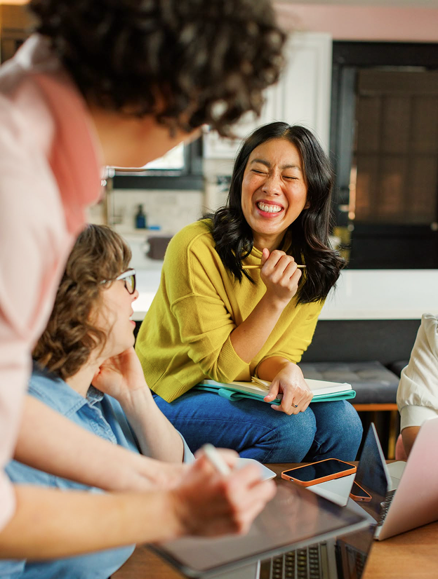 Three people engaged in a lively conversation, one woman in a yellow sweater laughing while holding a pencil and notebook.