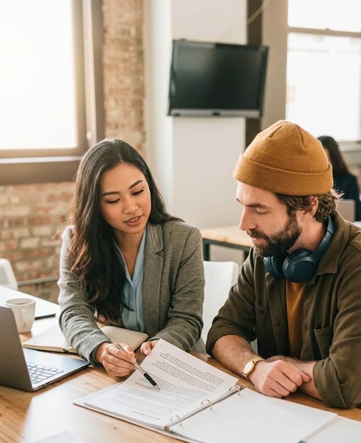 A corporate secretary in a grey blazer pointing with a pen at a document in a ring binder, explaining it to a client across a wooden desk in a naturally lit office. Korp.ph