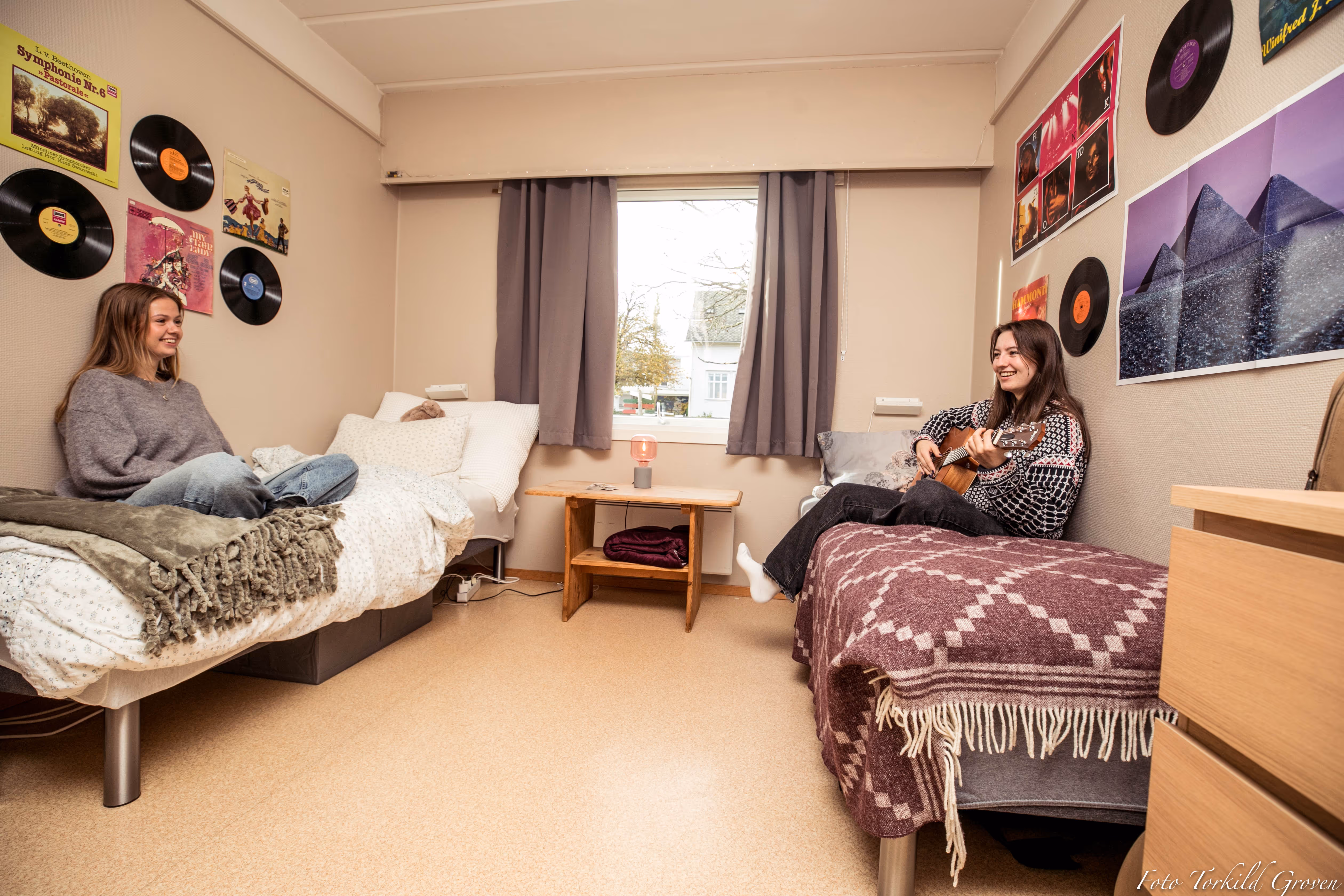To elever sitter på hvert sitt romseng ved Karmøy Folkehøgskole, den ene spiller gitar mens de prater og ler sammen i et personlig innredet rom med plakater og vinylplater på veggene