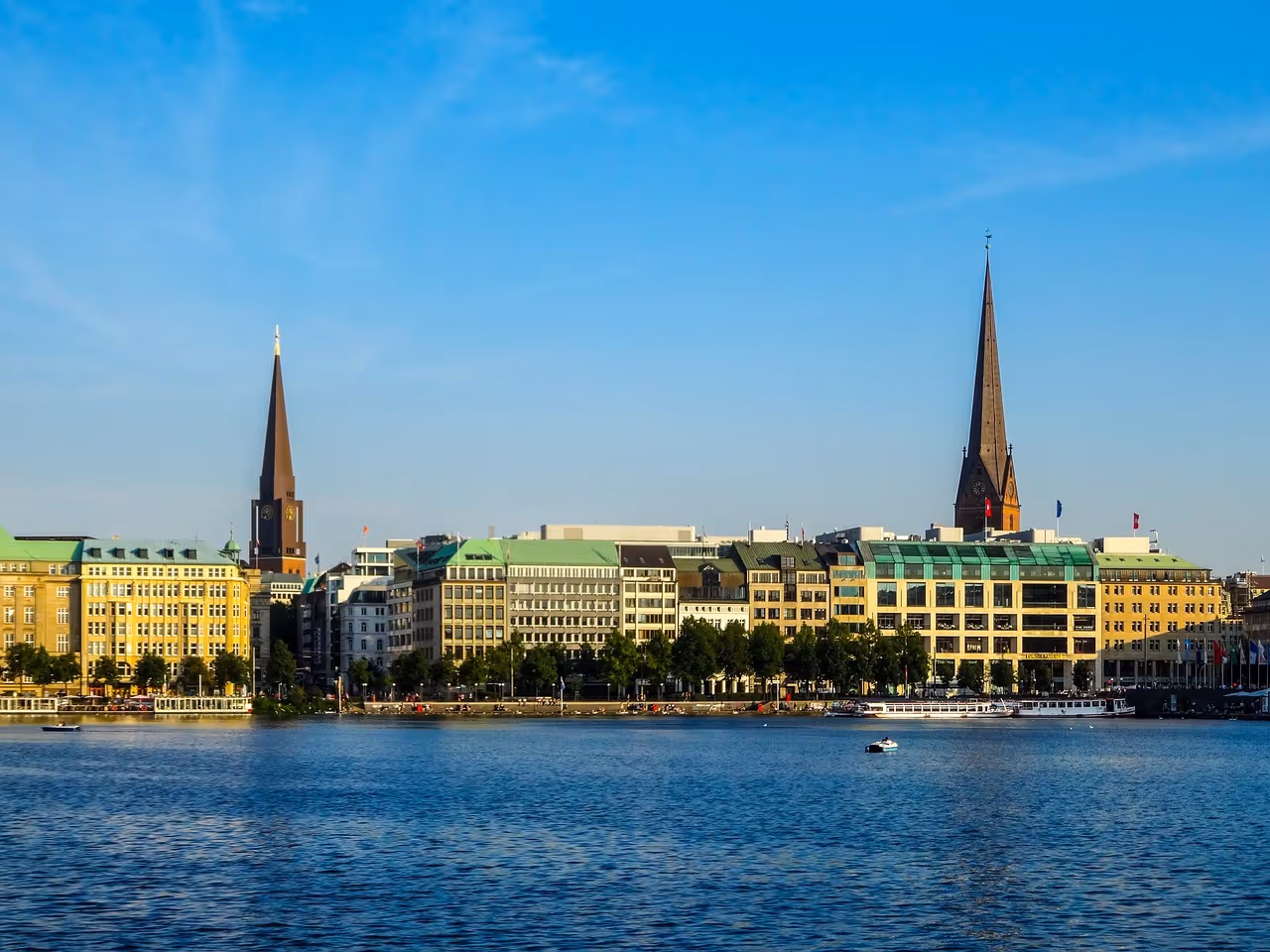 Blick auf die Binnenalster in Hamburg mit historischen Gebäuden und zwei hohen Kirchtürmen unter blauem Himmel.