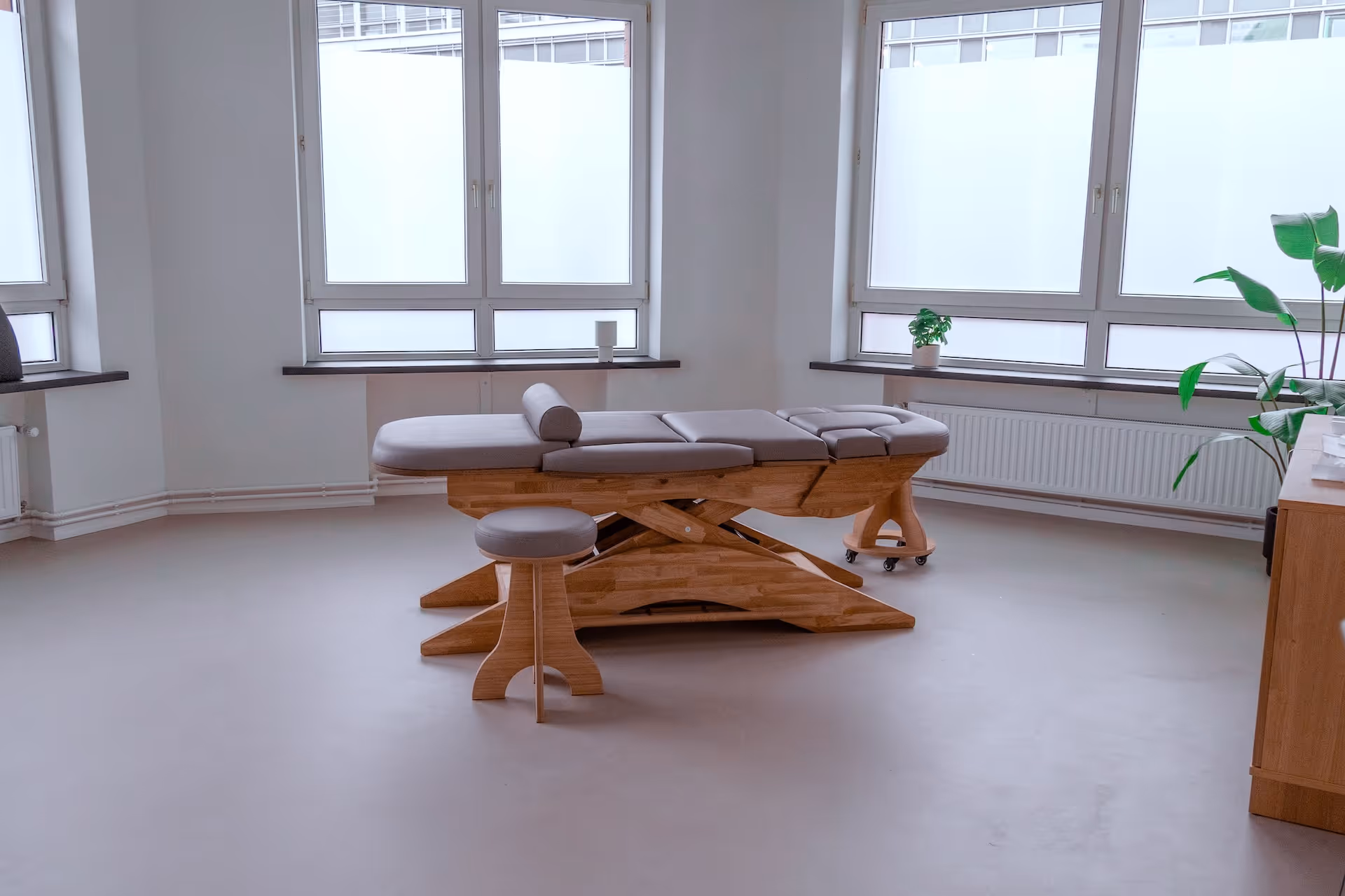 Helles Behandlungszimmer einer Osteopathie Praxis in Hamburg mit einem gepolsterten Massagetisch aus Holz und einem passenden Hocker vor großen Fenstern.