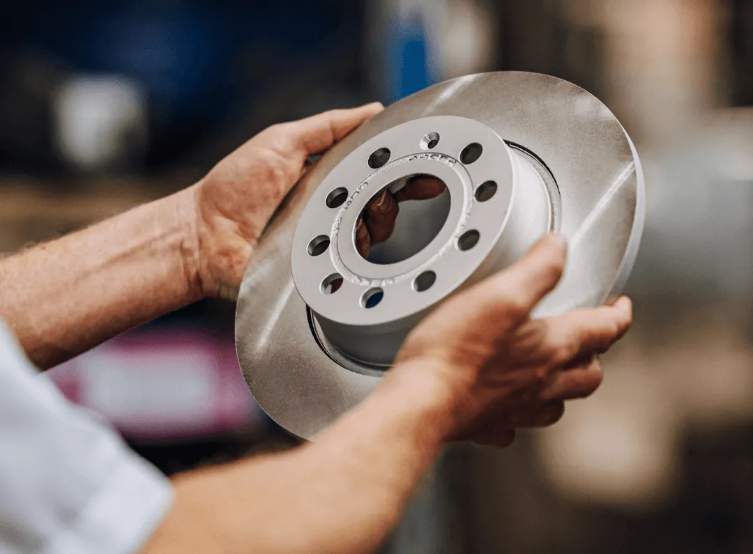 Hands holding a new silver brake disc rotor in a workshop setting.