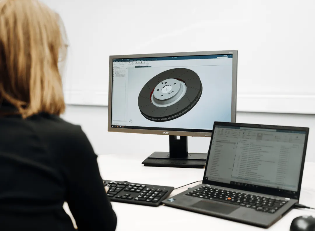 Person working at a desk with a desktop monitor displaying a 3D model of a brake disc and a laptop showing an email inbox.