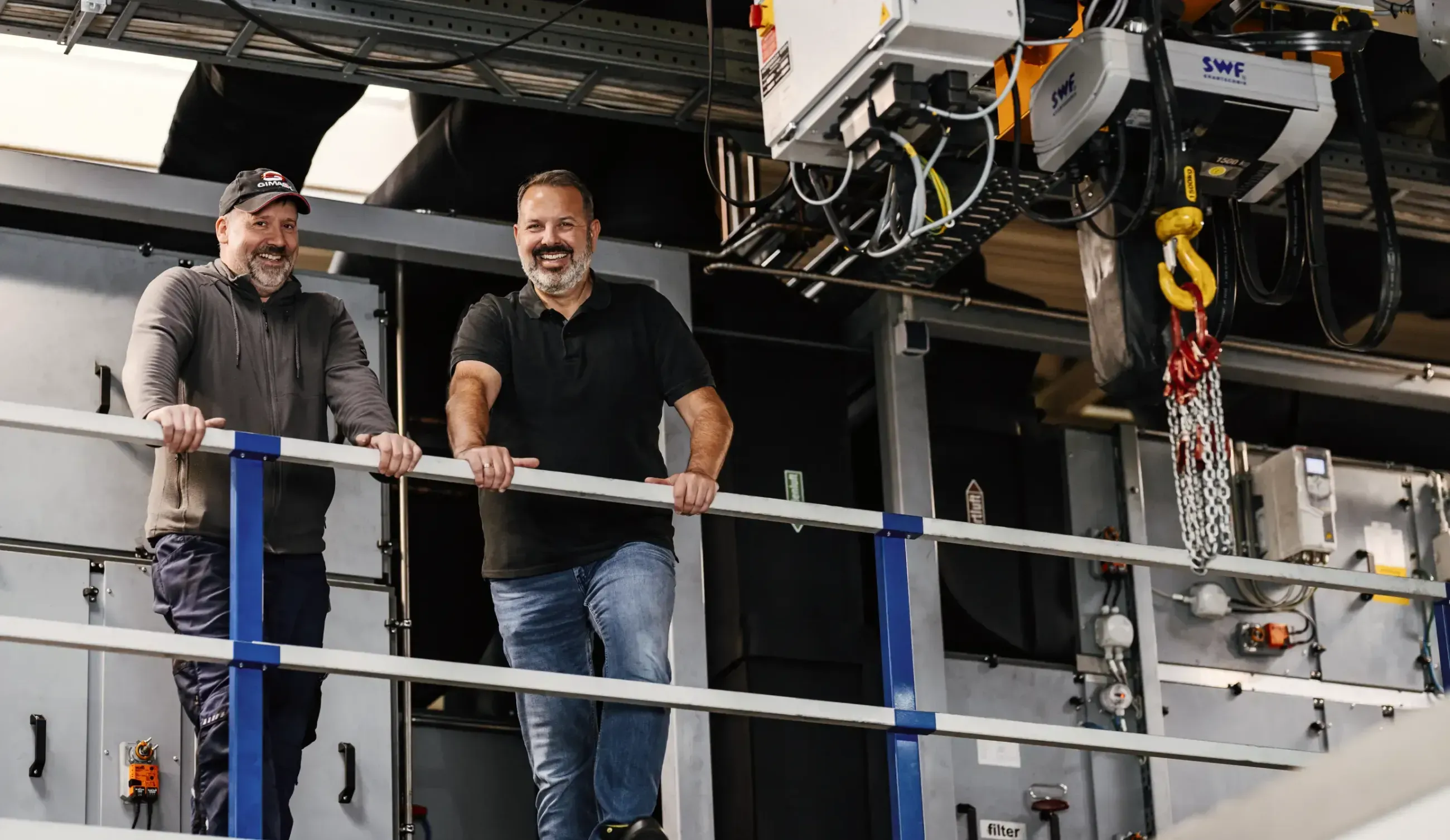 Two men smiling and standing behind a safety railing in an industrial facility with machinery overhead.