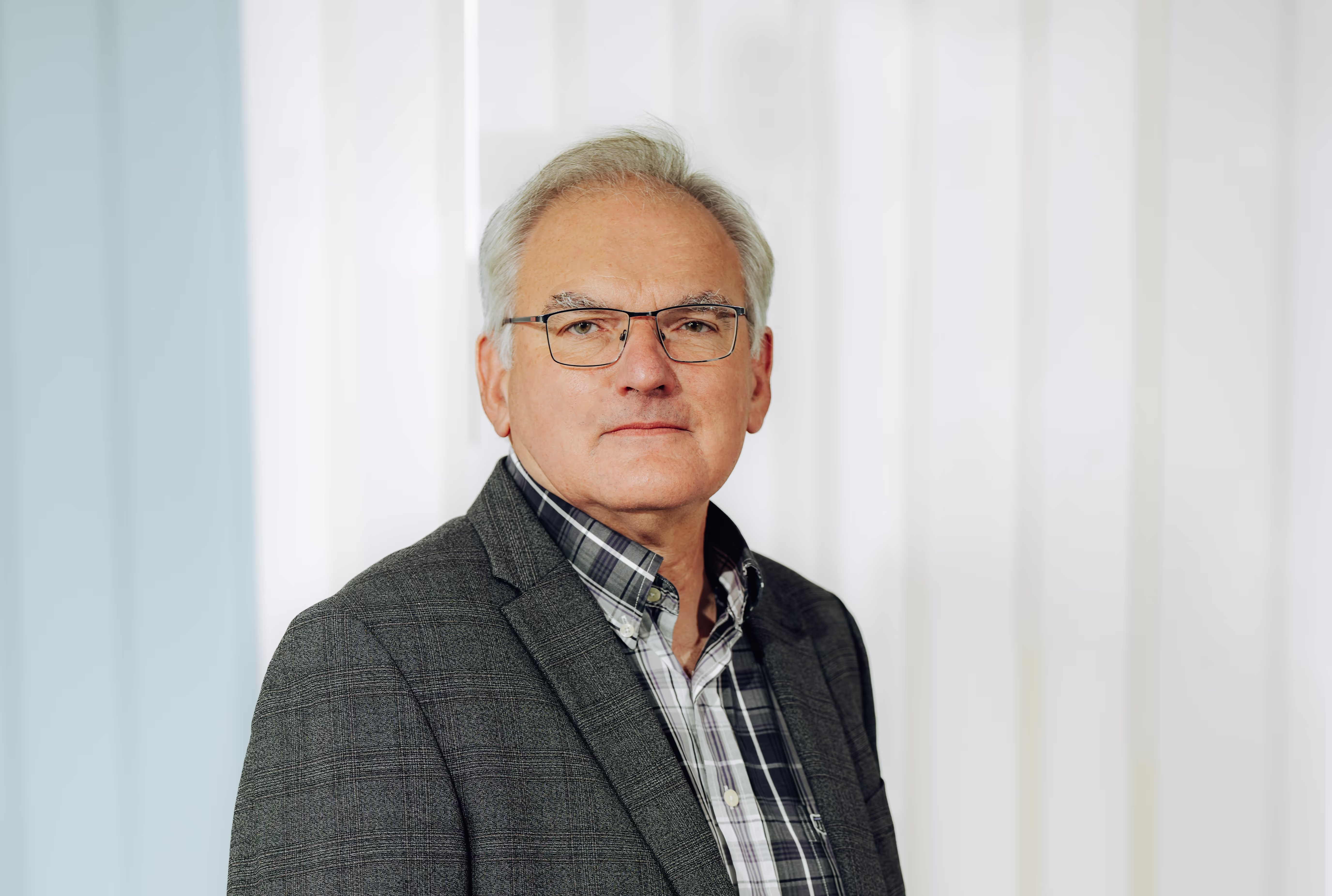 Middle-aged man with gray hair and glasses wearing a gray plaid blazer and checkered shirt against a light background.