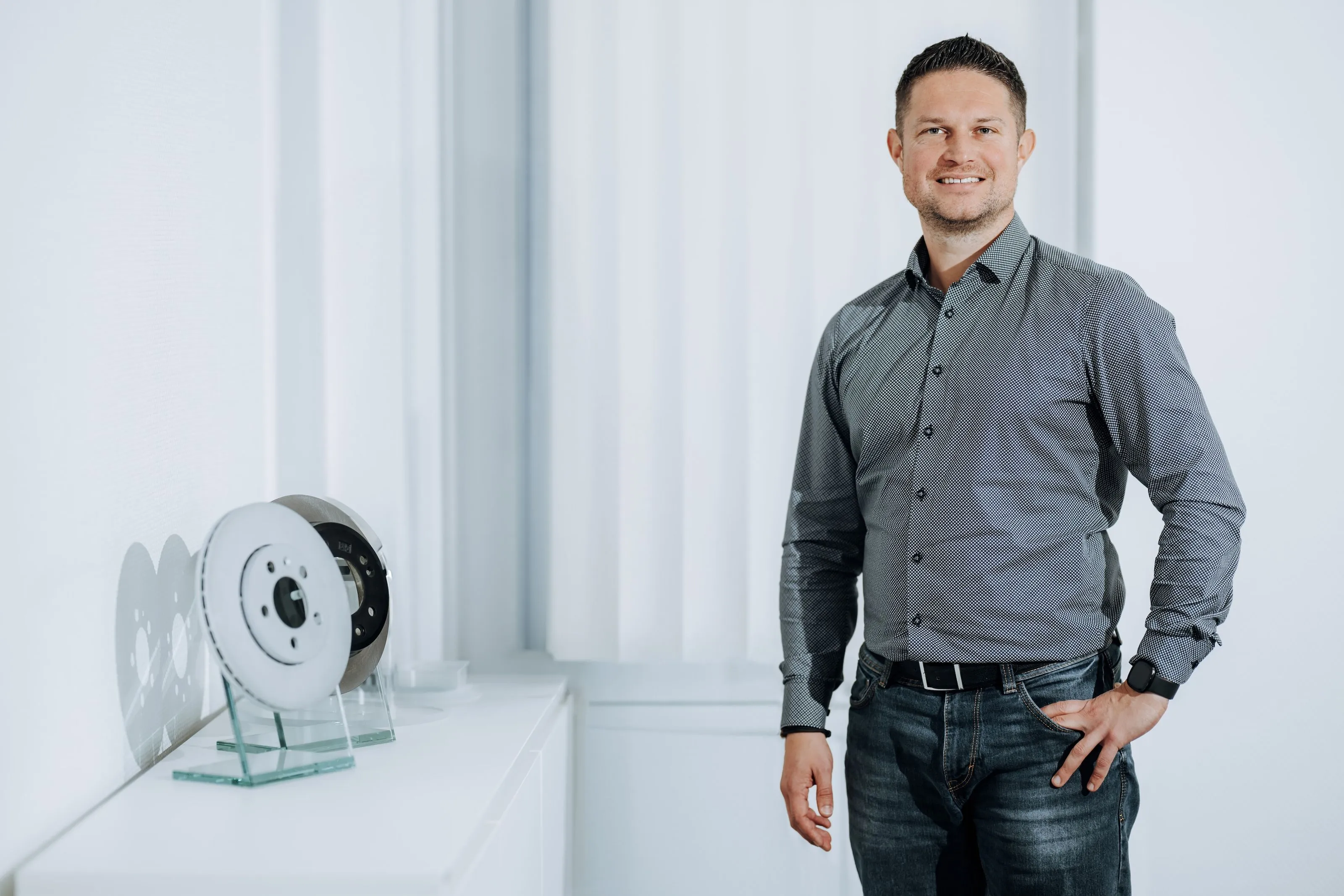 Man in a patterned button-up shirt and jeans standing next to a white table with displayed brake discs.