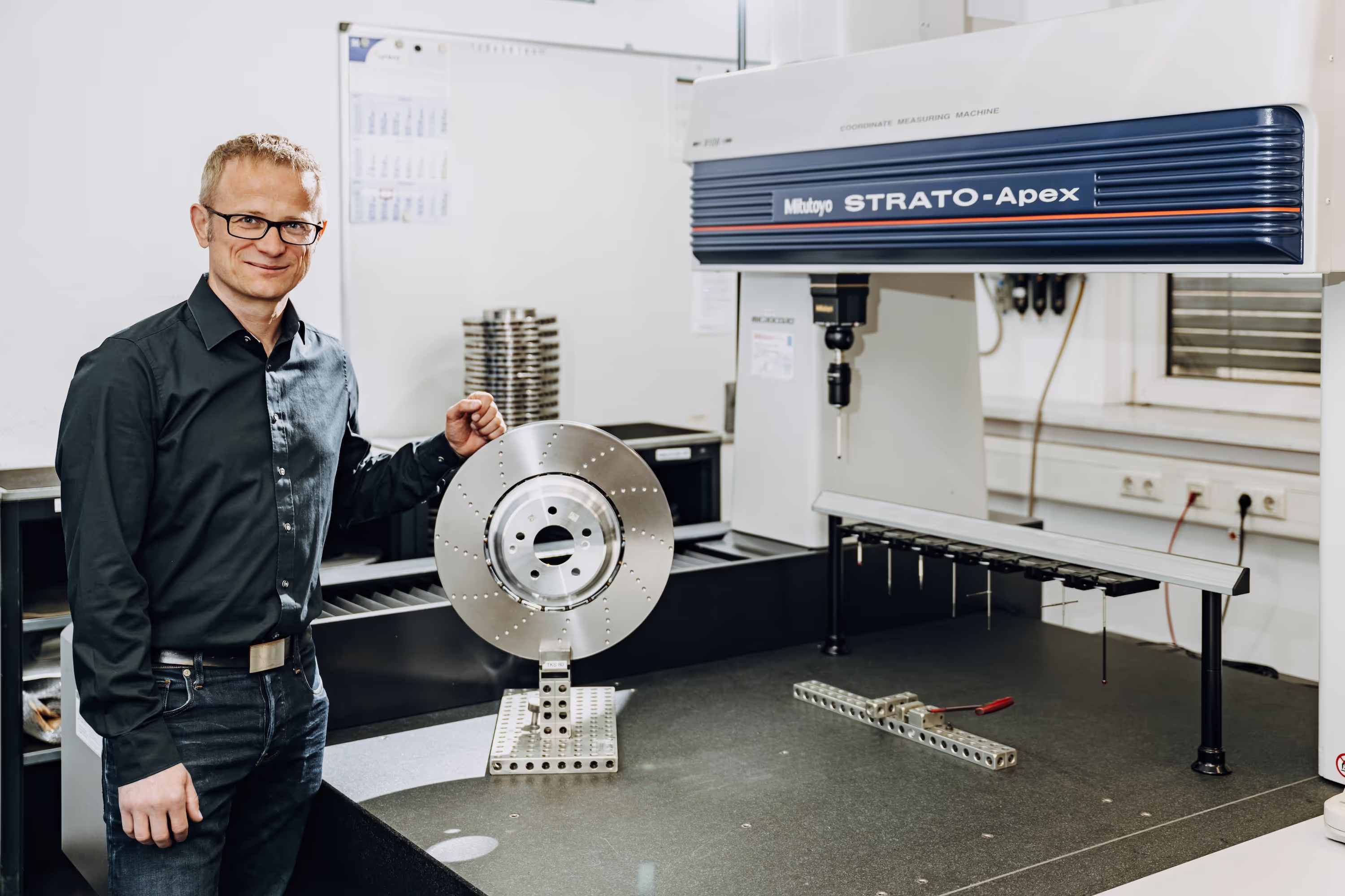 Man wearing glasses and a black shirt standing next to a metal disk mounted on a coordinate measuring machine in a workshop.