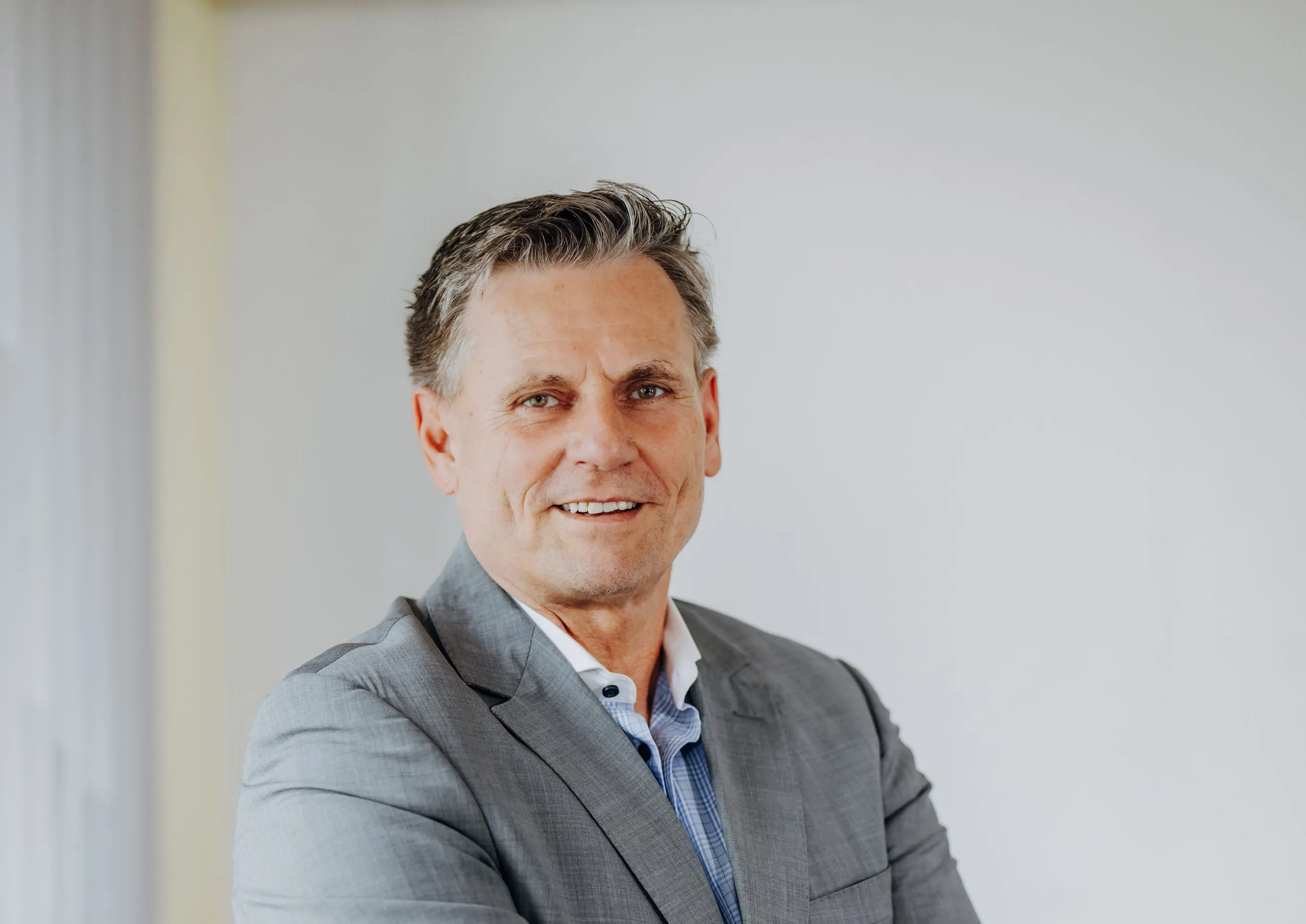 Smiling middle-aged man with gray hair wearing a gray suit and blue checkered shirt against a plain light background.