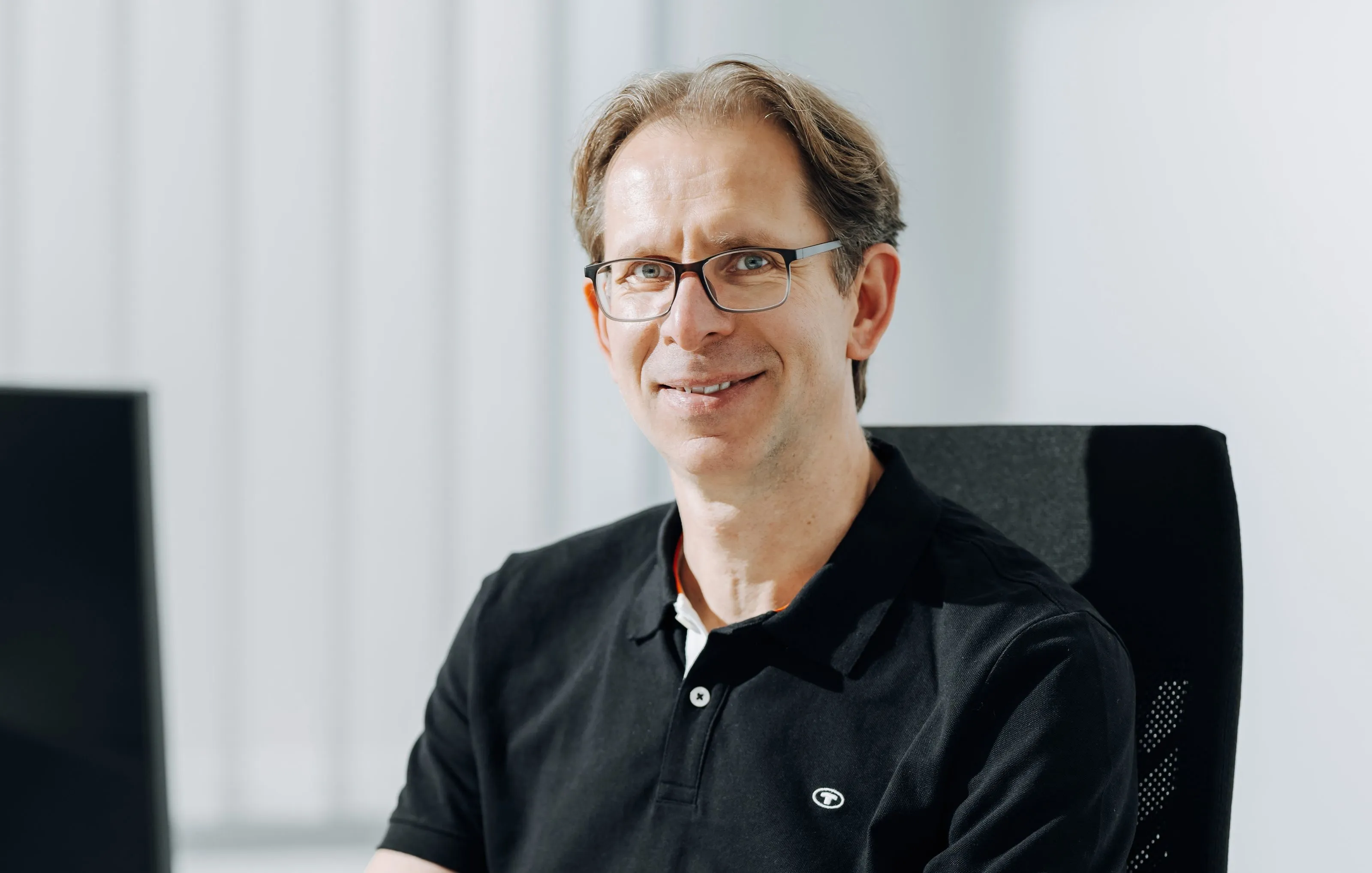 Smiling man with glasses wearing a black polo shirt sitting in an office chair.