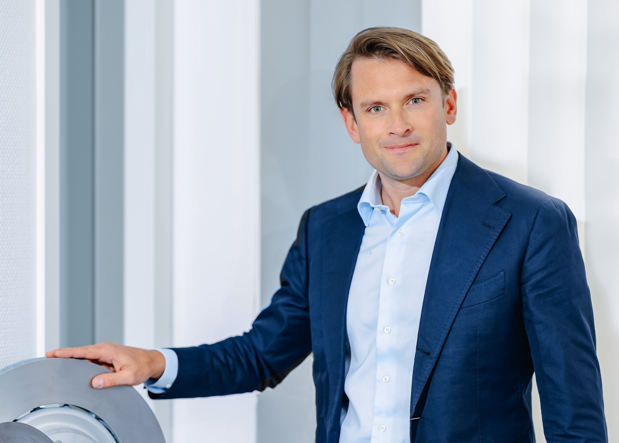 Man in a blue suit jacket and light blue shirt standing indoors, resting his hand on a round metallic object.