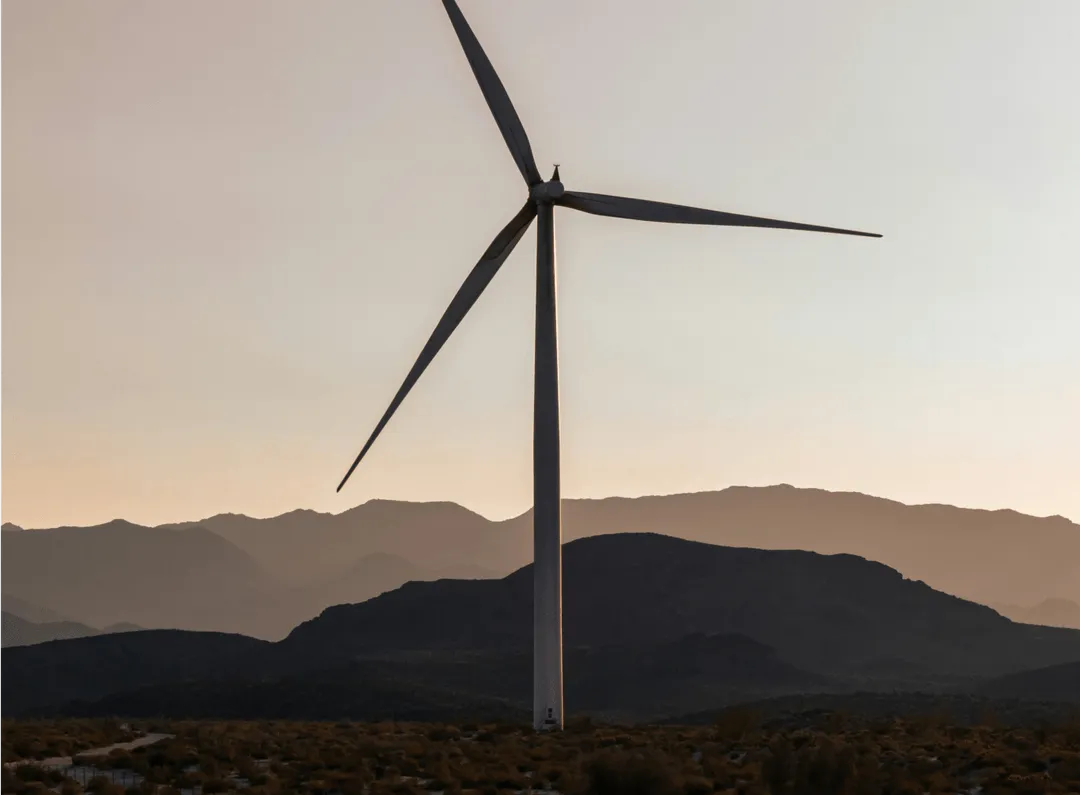 Single wind turbine standing in a desert landscape with mountains in the background at sunset.