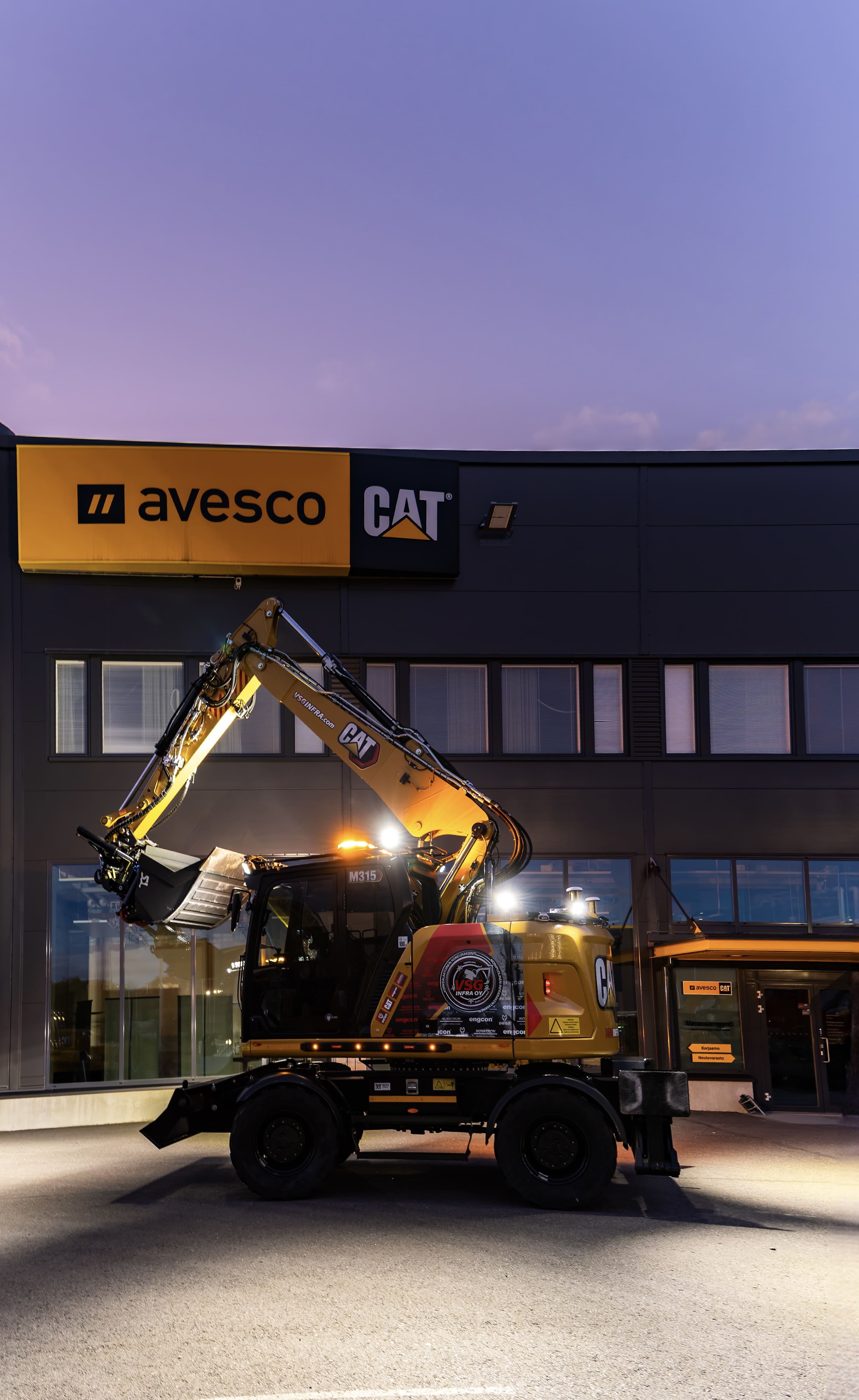 Keltainen CAT-kaivuri pysäköitynä rakennuksen eteen illalla valaistuna.