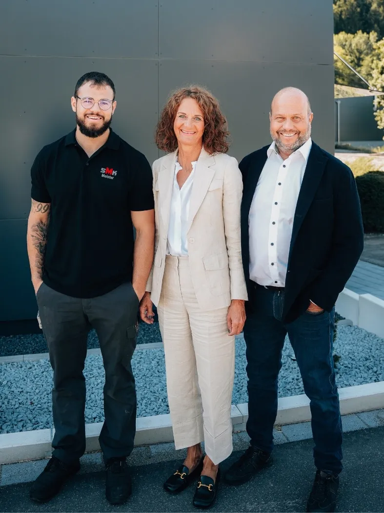 Drei lächelnde Personen stehen nebeneinander vor einer grauen Wand, zwei Männer in legerer Kleidung und eine Frau in einem hellen Anzug.