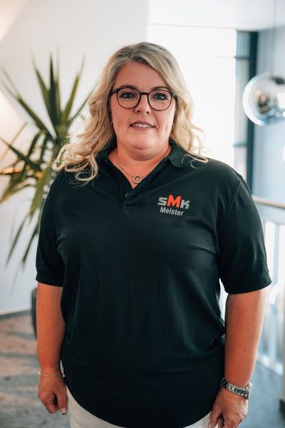 Frau mit blonden Haaren, Brille und schwarzem Polohemd mit SMK Meister-Logo steht in einem modernen Innenraum mit Pflanzen im Hintergrund.
