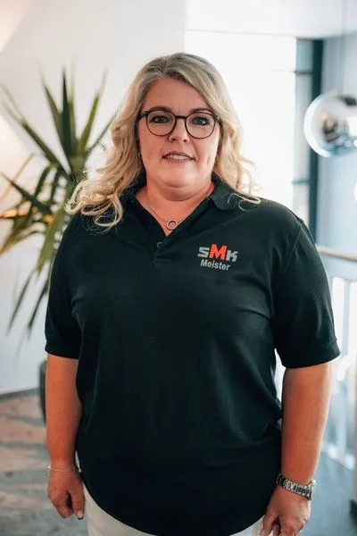 Frau mit blonden Haaren, Brille und schwarzem Polohemd mit SMK Meister-Logo steht in einem modernen Innenraum mit Pflanzen im Hintergrund.