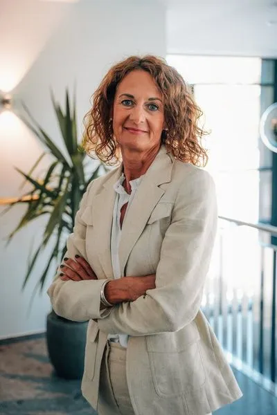 Geschäftsfrau mit lockigem Haar und beigem Anzug steht mit verschränkten Armen in einem modernen Büro.