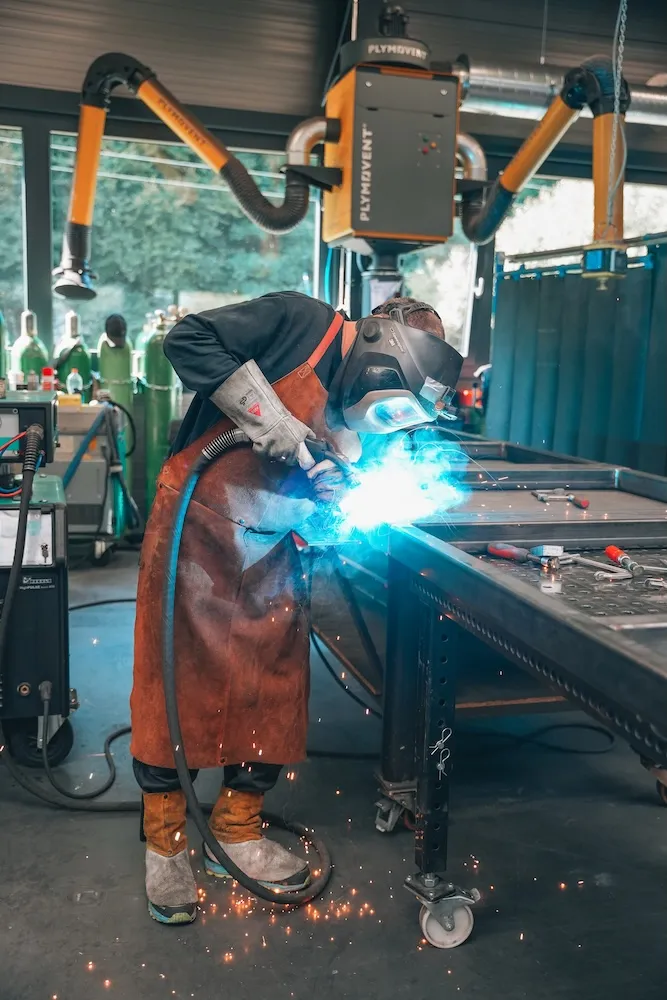 Schweißer mit Schutzhelm und Lederschürze schweißt an einem Metalltisch in einer Werkstatt, Funken fliegen.