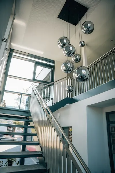Moderne Treppe mit Edelstahlgeländer in einem hellen Raum, darüber hängende reflektierende Kugelpendelleuchten.