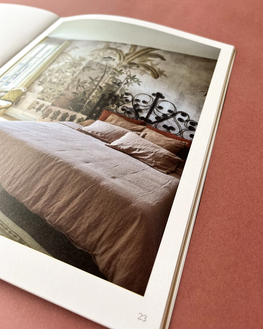 Photo représentant une page de catalogue montrant un lit double avec une couette et des oreillers en lin brun clair. La tête de lit est en métal noir avec des motifs décoratifs en spirale. Le mur derrière le lit présente une fresque murale représentant des plantes et un palmier. À gauche, une fenêtre laisse entrer la lumière naturelle et un petit fauteuil est visible. La page est numérotée « 23 ».