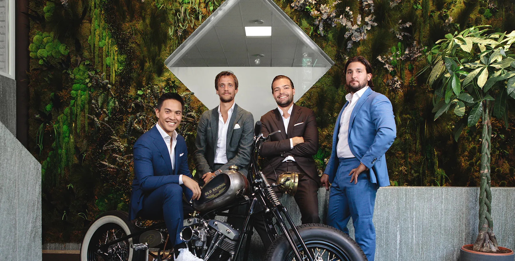 Un groupe d'hommes en costumes posant près d'une moto vintage dans un espace moderne avec un mur végétal en arrière-plan.