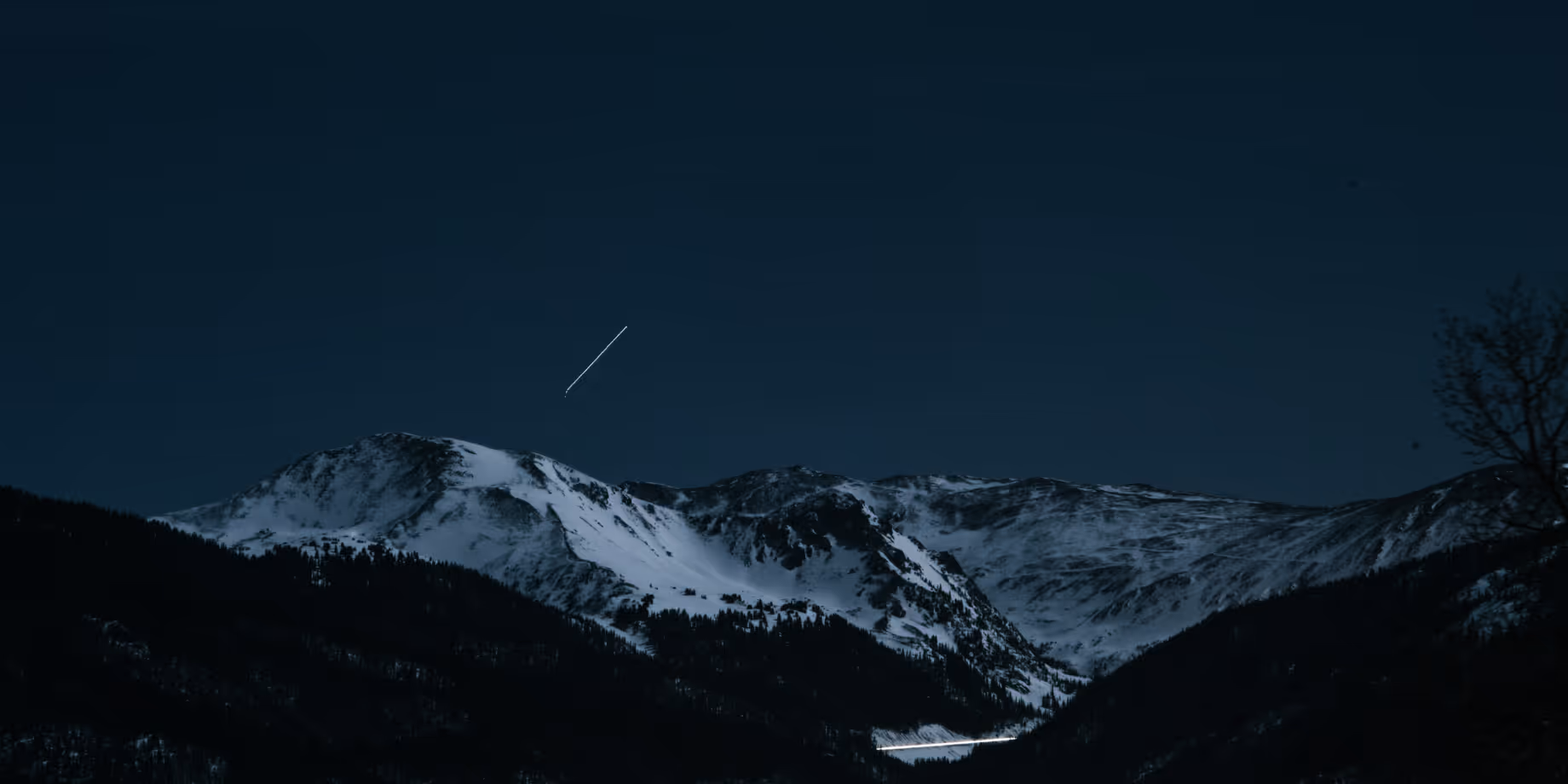Snow-covered mountain range under a dark night sky with a visible streak of a shooting star or airplane light.