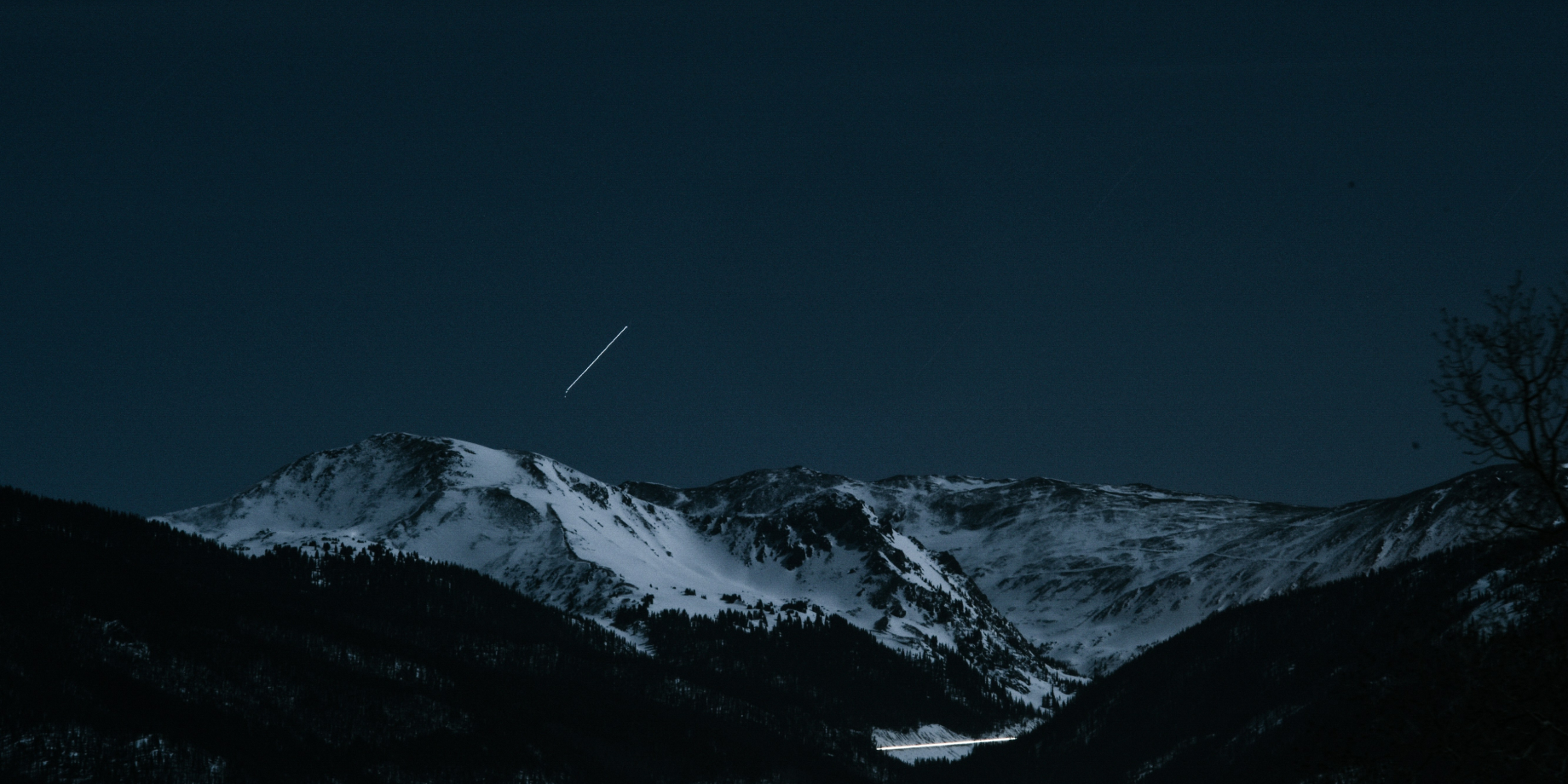 Snow-covered mountain range under a dark night sky with a visible streak of a shooting star or airplane light.