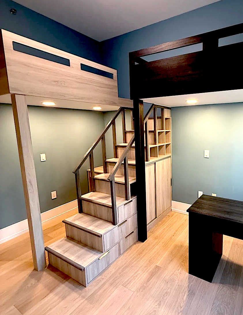 Modern room with wooden loft beds connected by a staircase with built-in storage and a dark wooden desk.