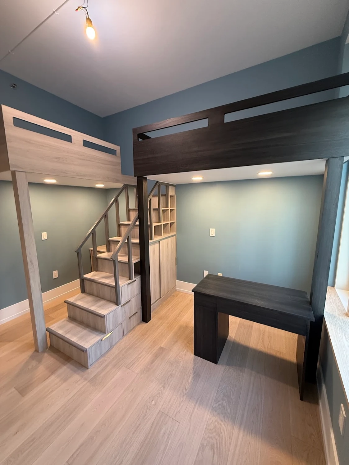 Modern bedroom with light wooden loft bed featuring stairs and built-in storage, and a dark wooden desk underneath another loft bed.