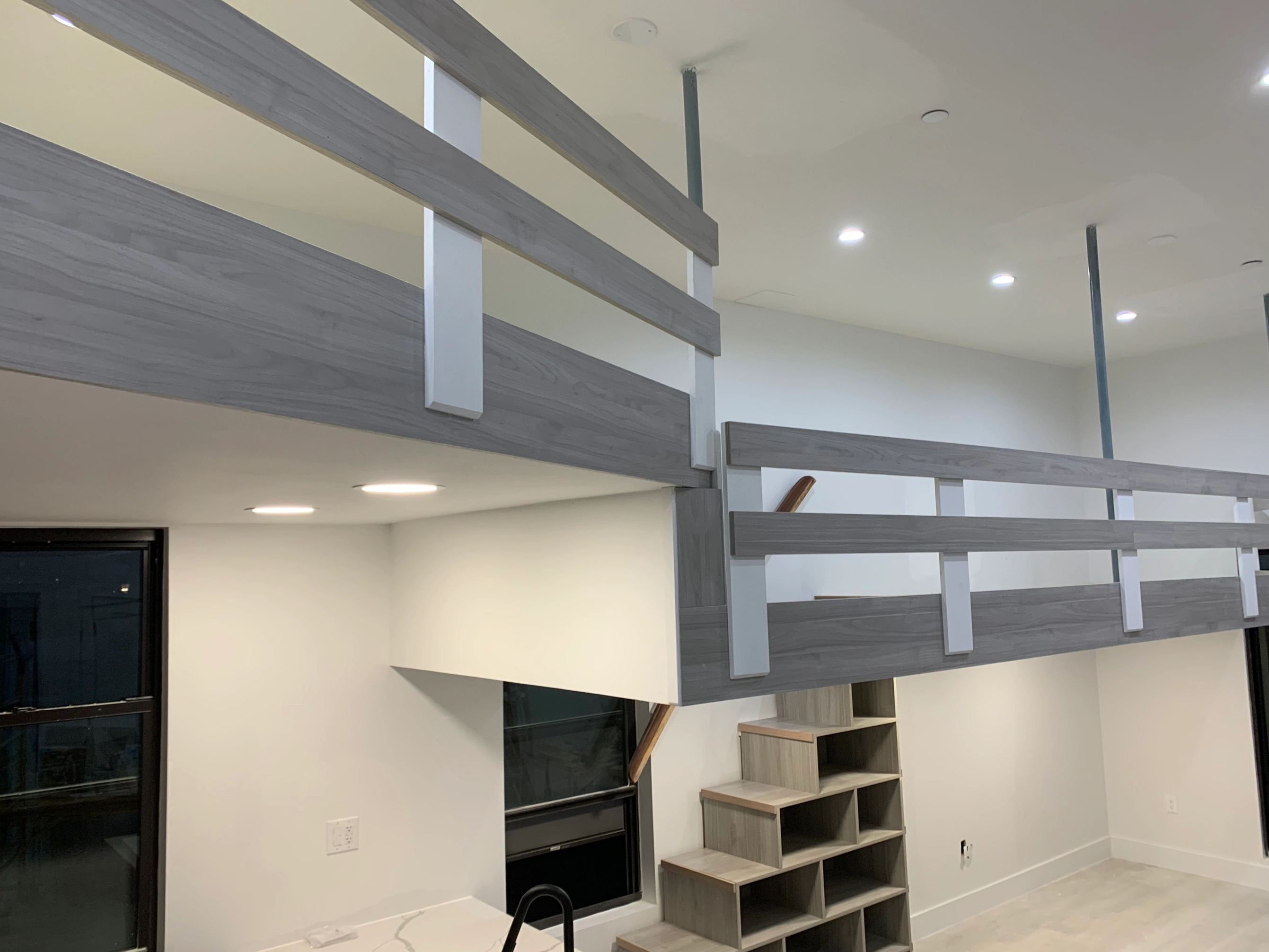 Indoor loft area with gray wooden railings, built-in wooden staircase with storage cubbies, and white walls under ceiling lights.