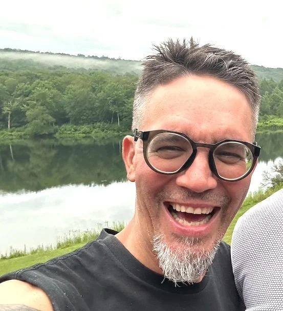 Smiling man with glasses and a salt-and-pepper beard outdoors near a calm lake and forest.