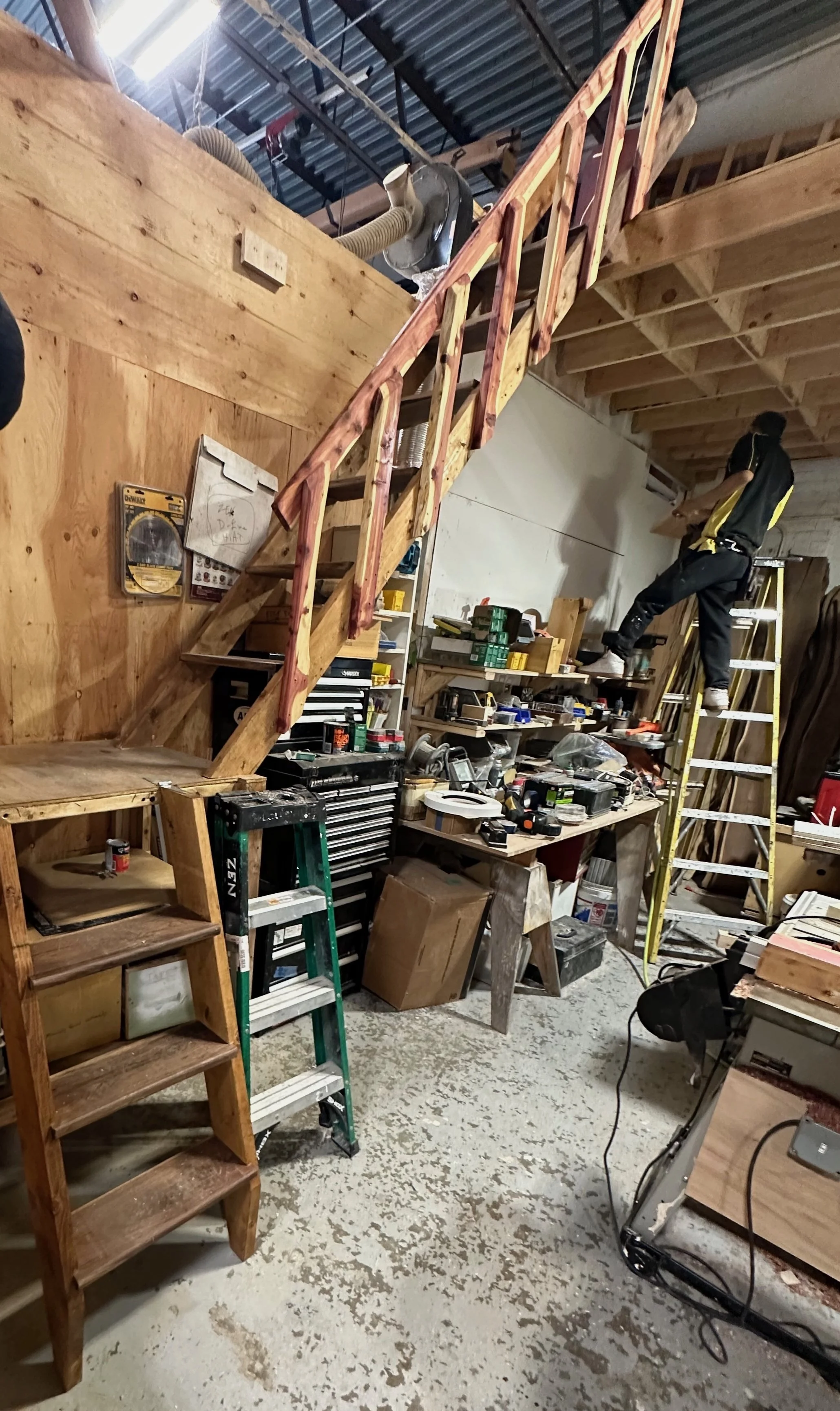 A workshop with plywood walls, wooden staircase, shelves filled with tools and supplies, and a man standing on a yellow ladder working near the ceiling.
