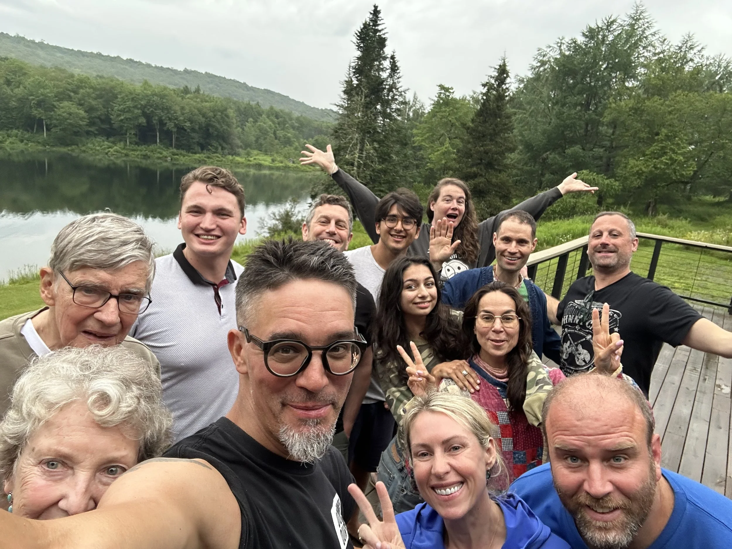 Group of smiling people posing for a selfie outdoors near a lake and forested hills on a cloudy day.