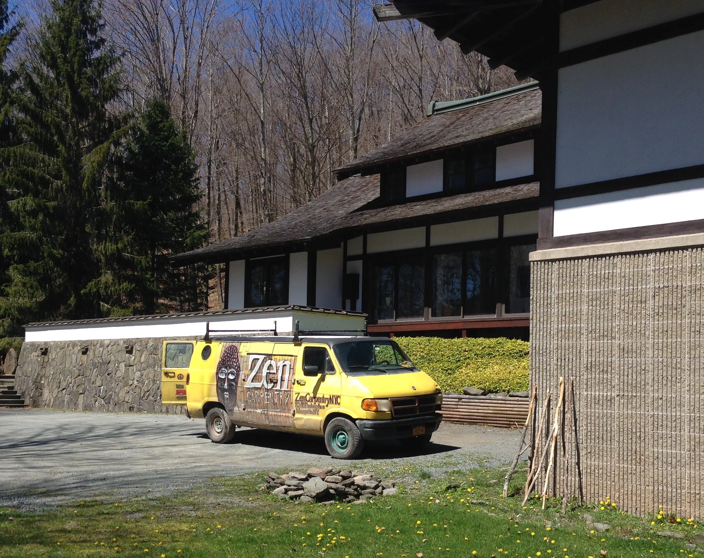 Yellow Zen Carpentry van parked on gravel near a Japanese-style building with a stone wall and leafless trees in the background.