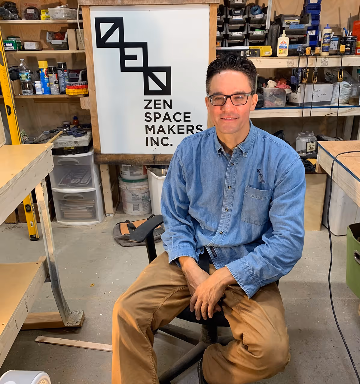 founder of of Zen space makers wearing glasses, denim shirt, and khaki pants sitting on a chair in a workshop with a Zen Space Makers Inc. sign behind him.