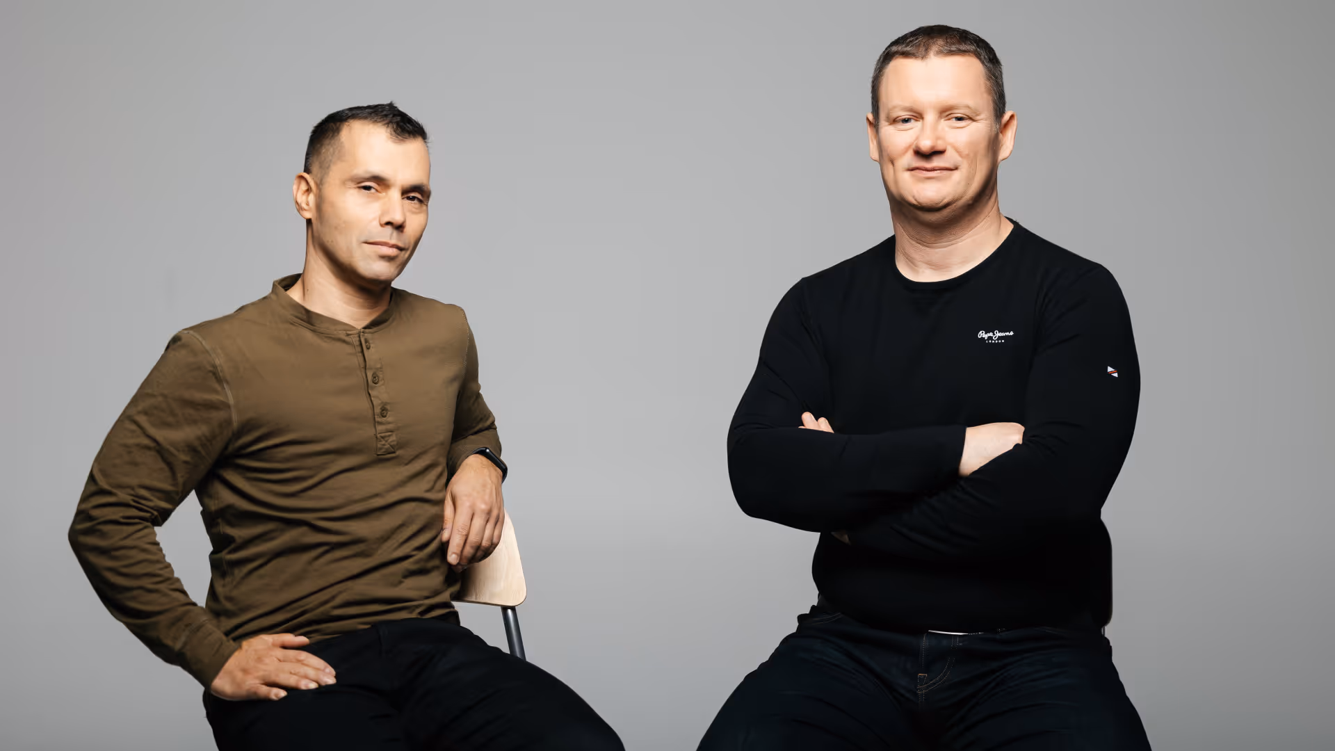 Egy barna és fekete pólós férfi bárszéken ülve pózol egy szürke háttér előtt