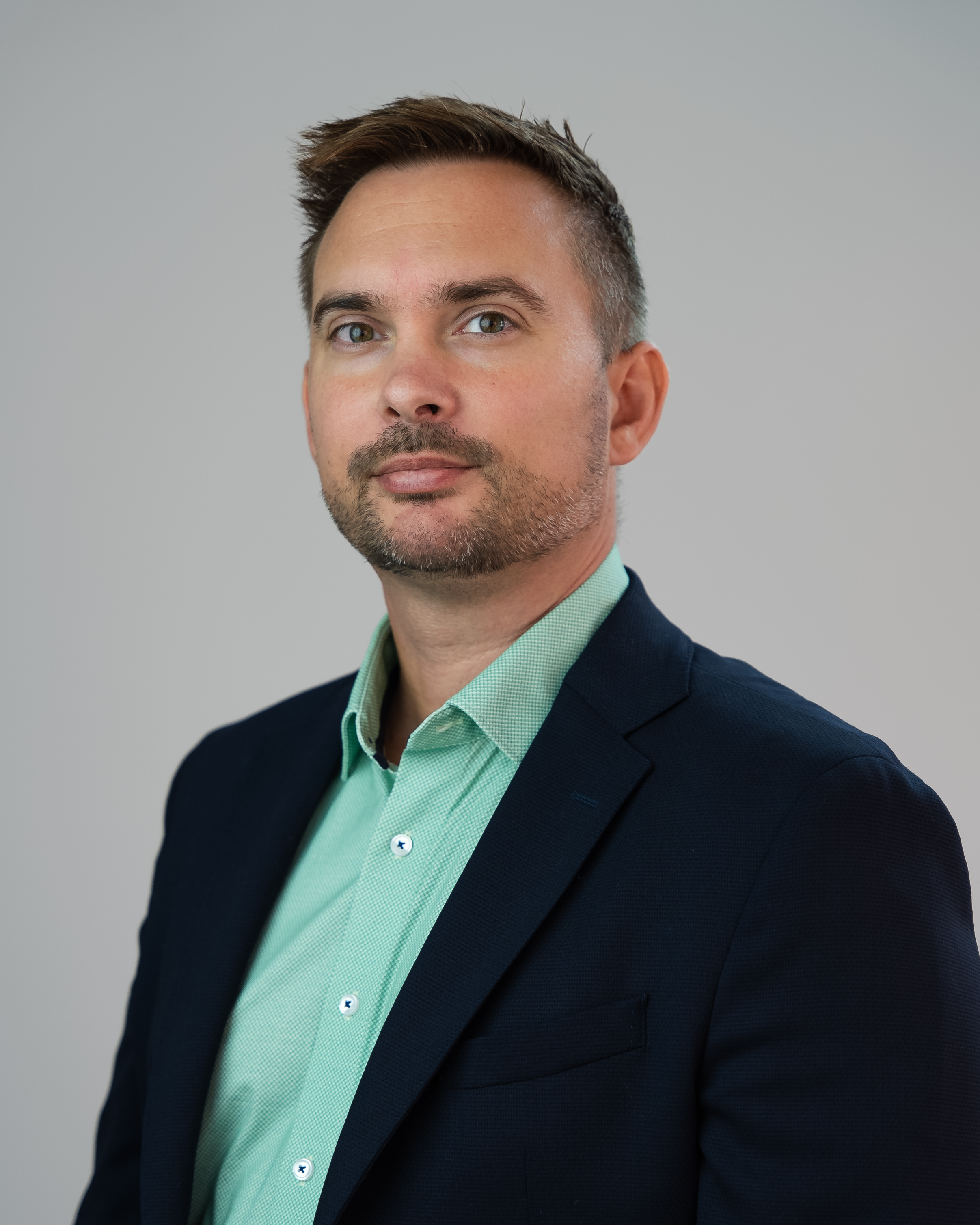 Szakállas fiatal férfi sötét zakóban, zöld ingben világos háttér előtt