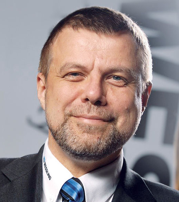 Borostás középkorú férfi öltönyben világos, szürke feliratos háttér előtt