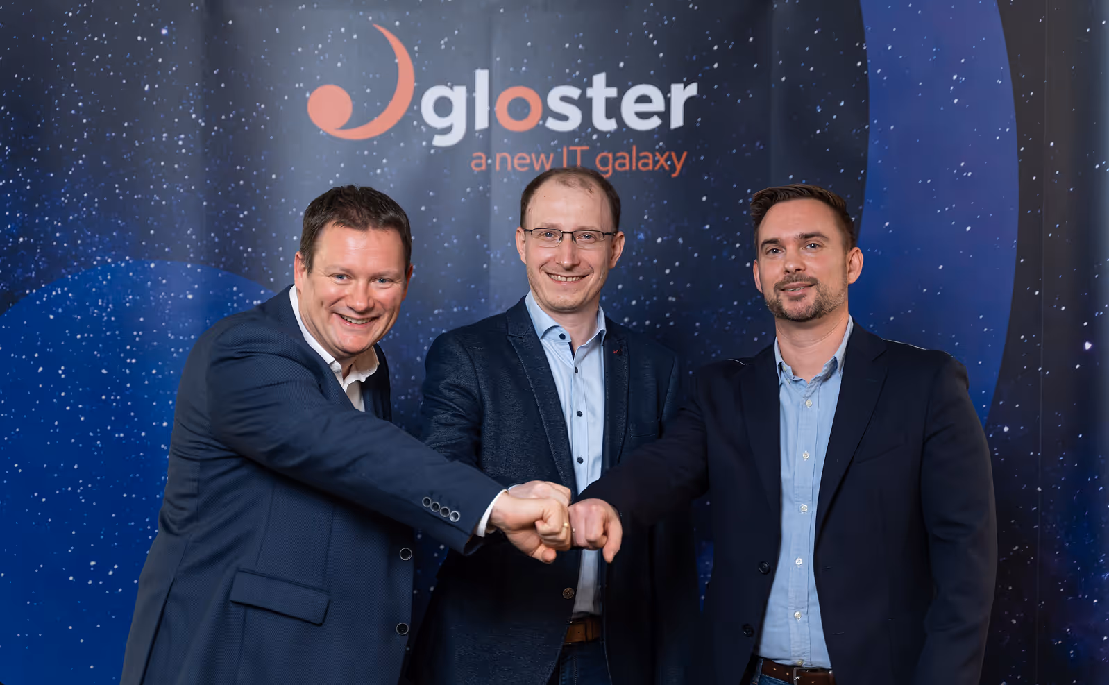 3 férfi öltönyben az ökleiket középre tartva egy Gloster design fal előtt