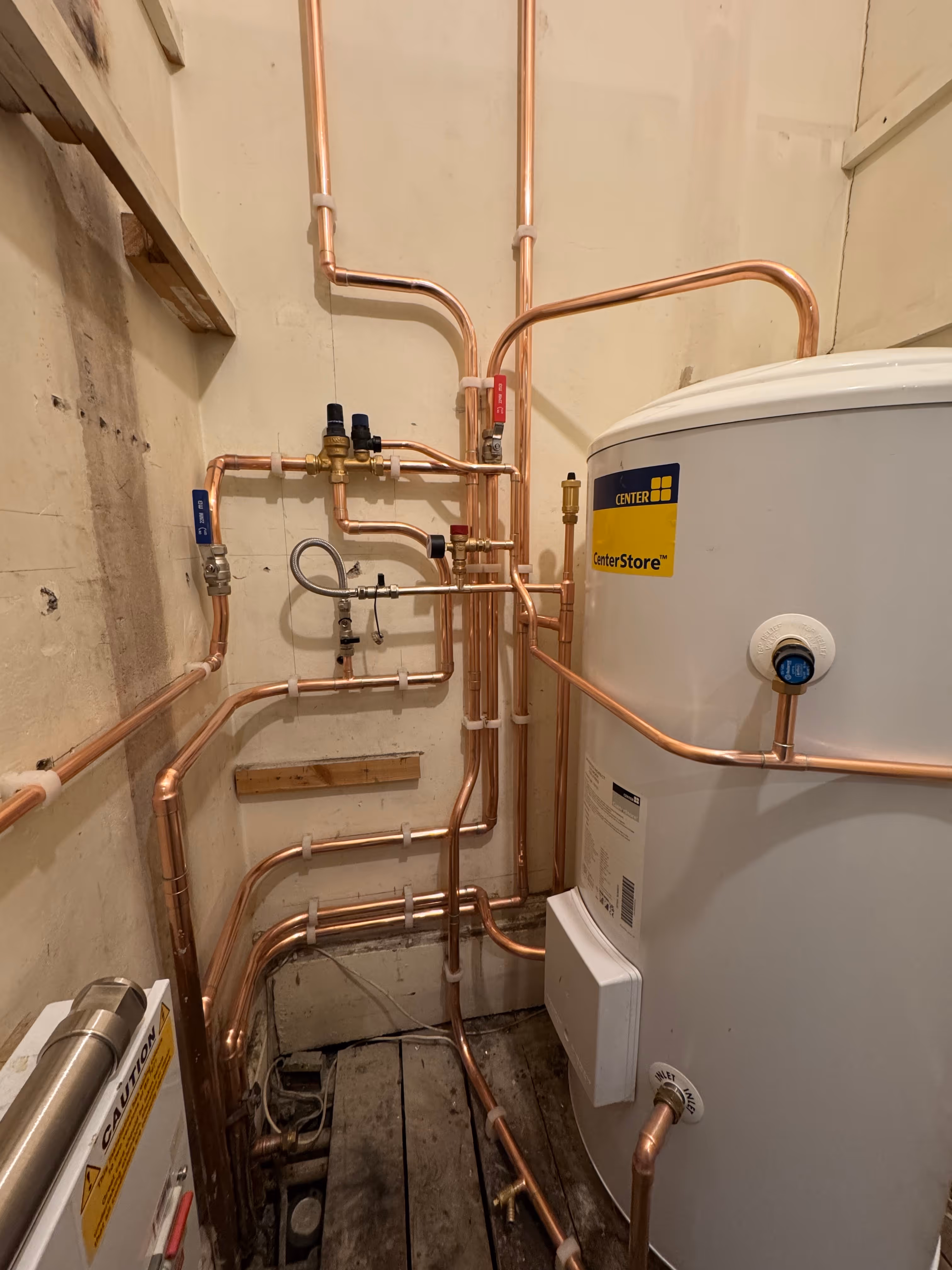 Copper piping system installed on a beige wall connected to a large white CenterStore water heater tank inside a utility room with wooden floorboards.