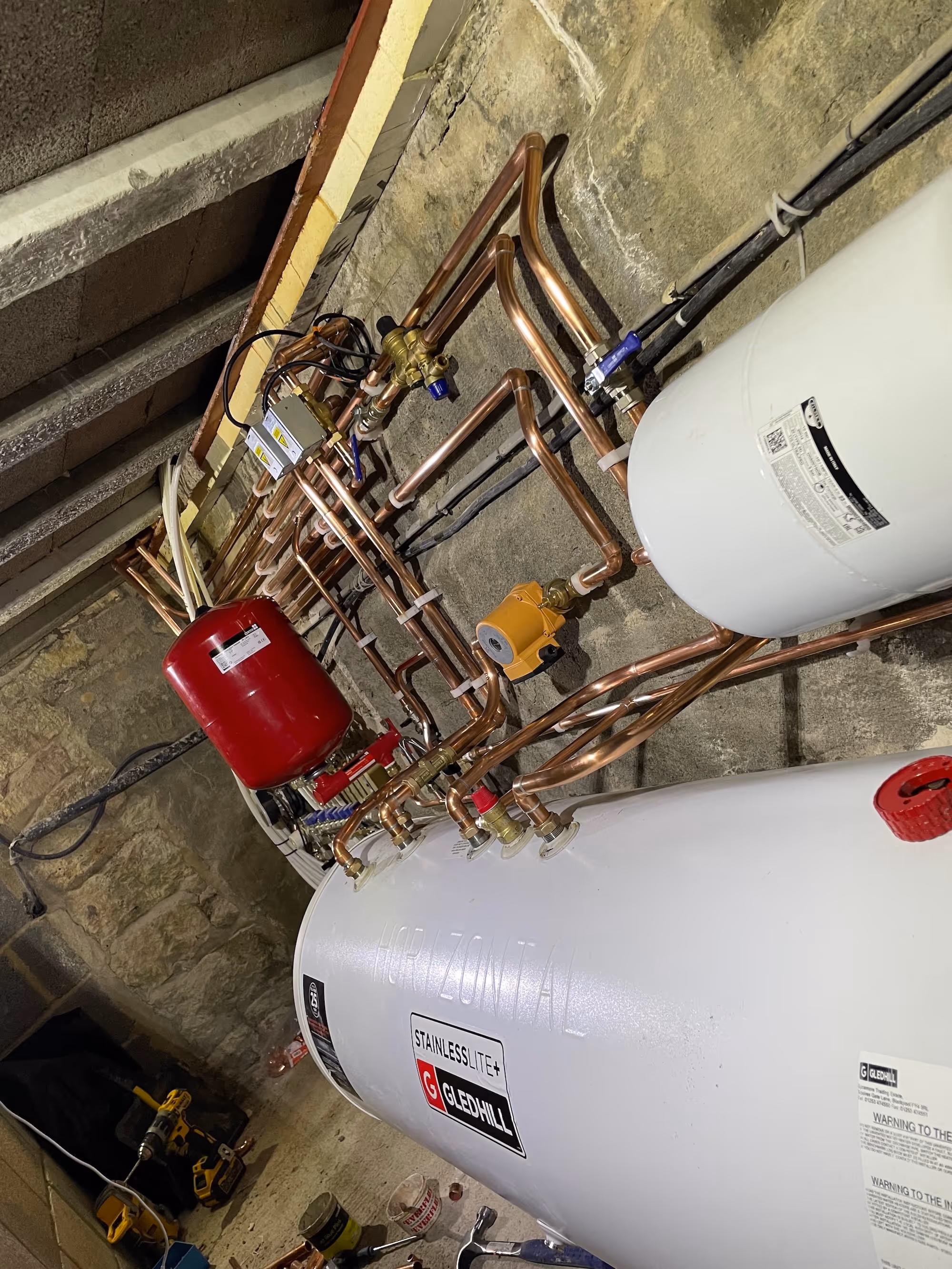 Horizontal Gledhill Stainless Lite+ water heater with connected copper piping and red expansion tank in a basement with stone walls and work tools on the floor.