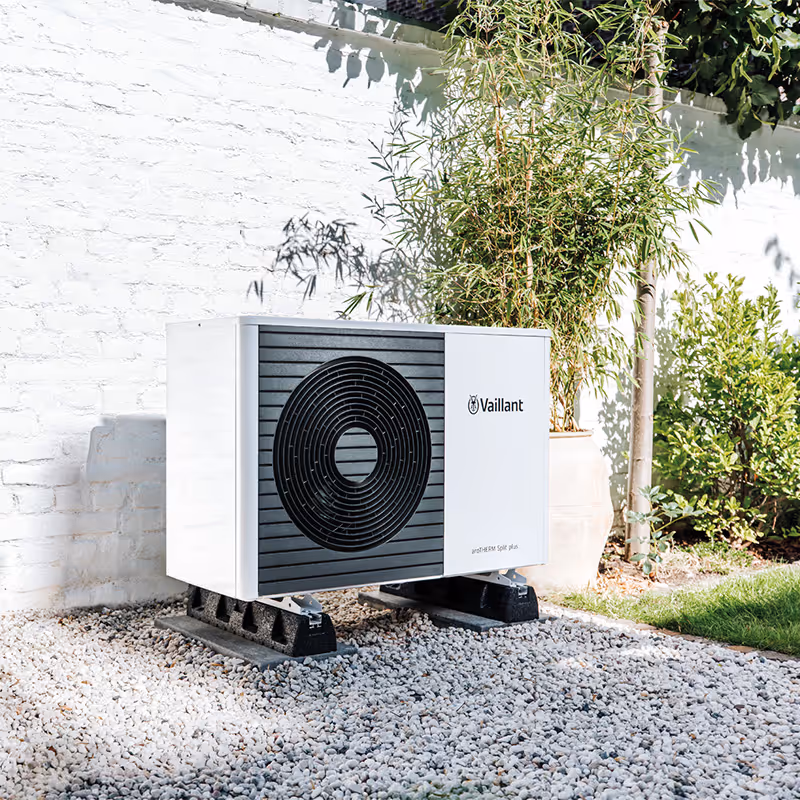Outdoor Vaillant aroTHERM Split plus heat pump unit installed on gravel with plants and white brick wall background.