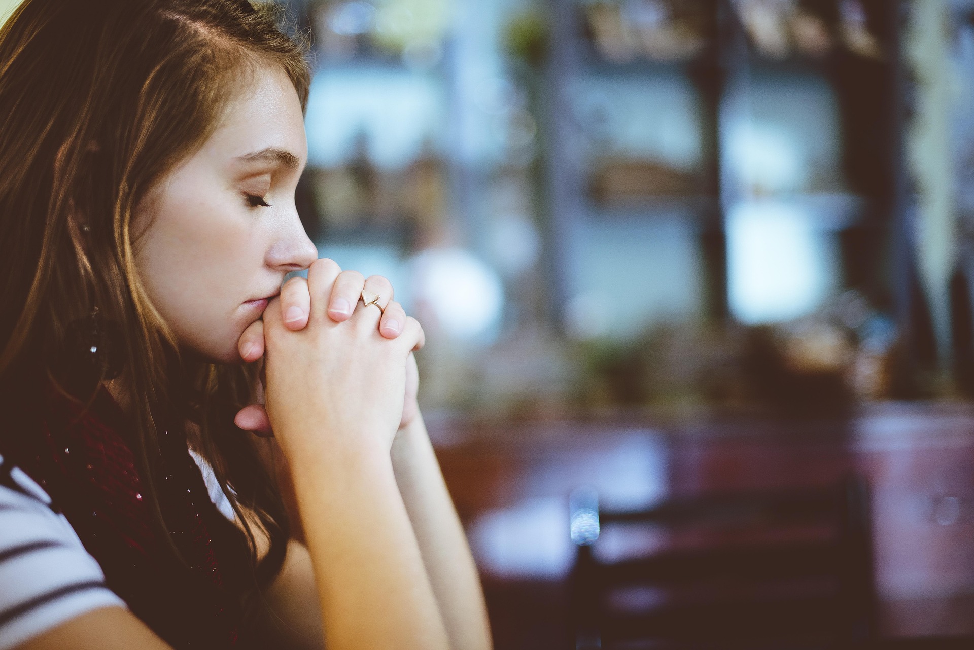 Young woman with closed eyes resting her chin on clasped hands in a blurred indoor setting.