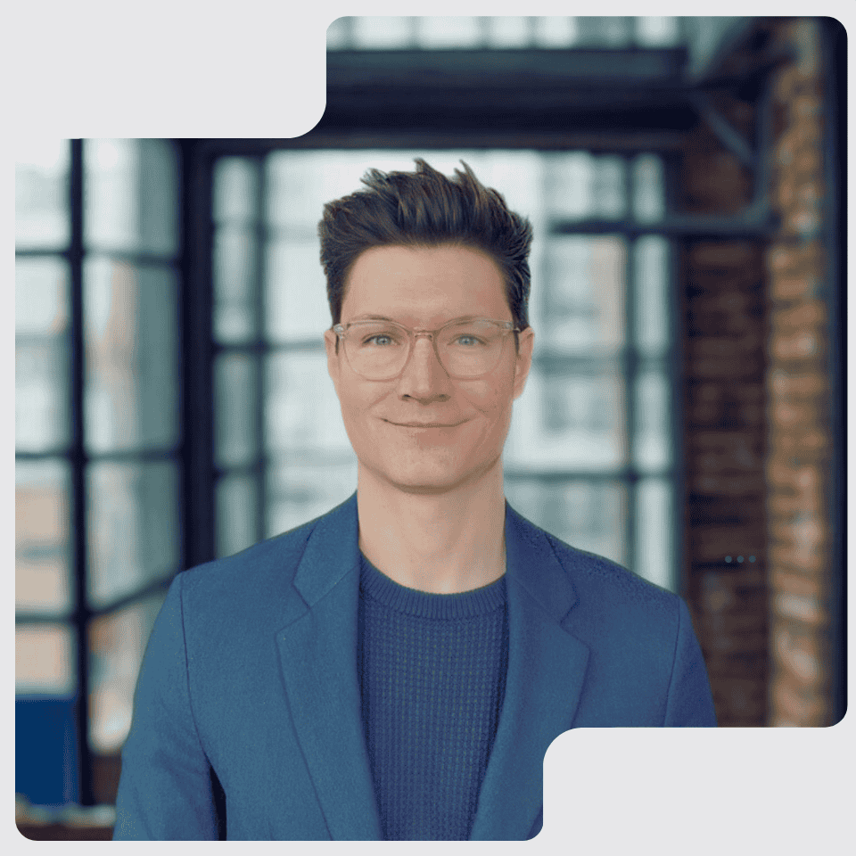 Smiling man with glasses wearing a blue blazer and sweater in a modern office setting.