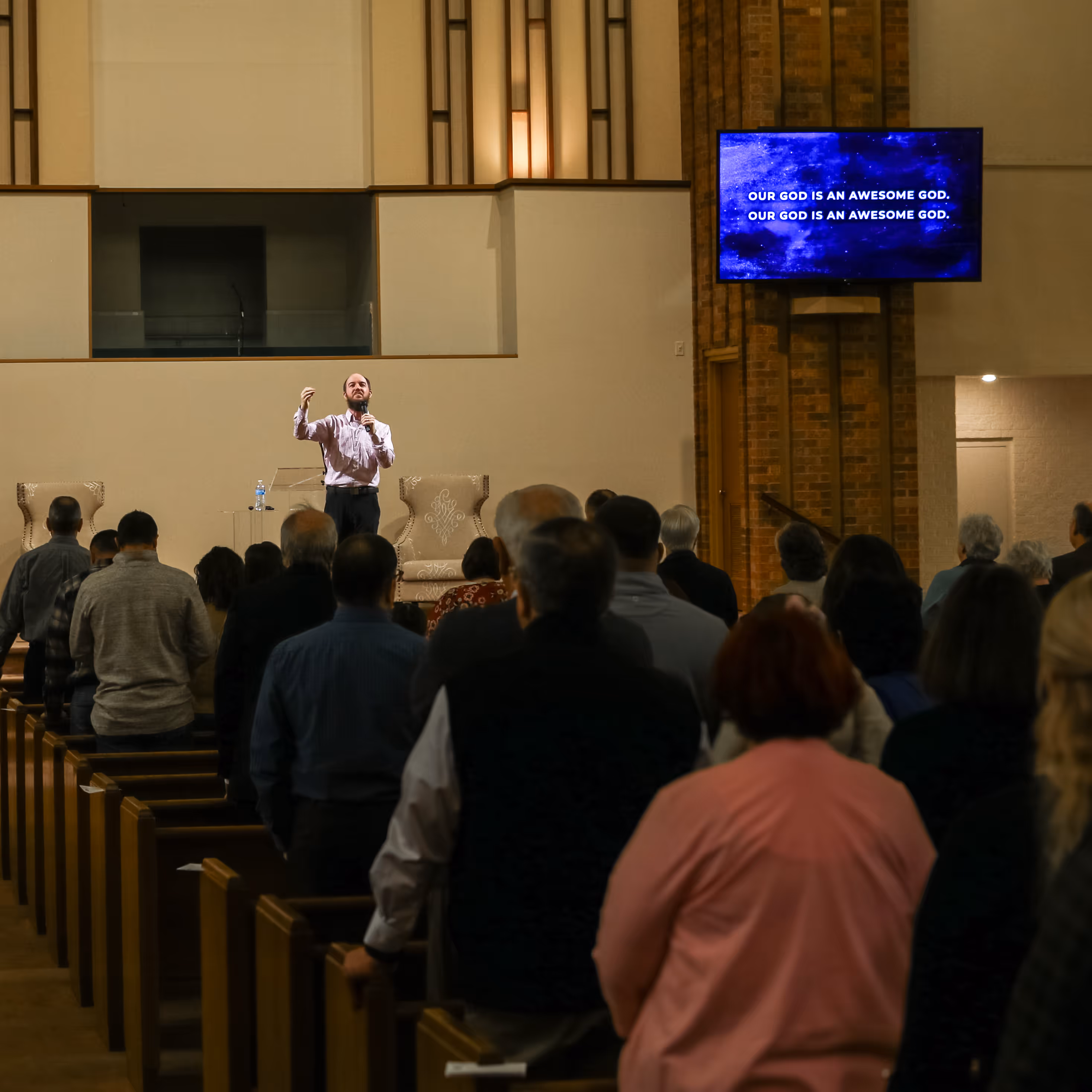 Man singing in front of a standing congregation inside a church, with a screen showing the text 'OUR GOD IS AN AWESOME GOD.'