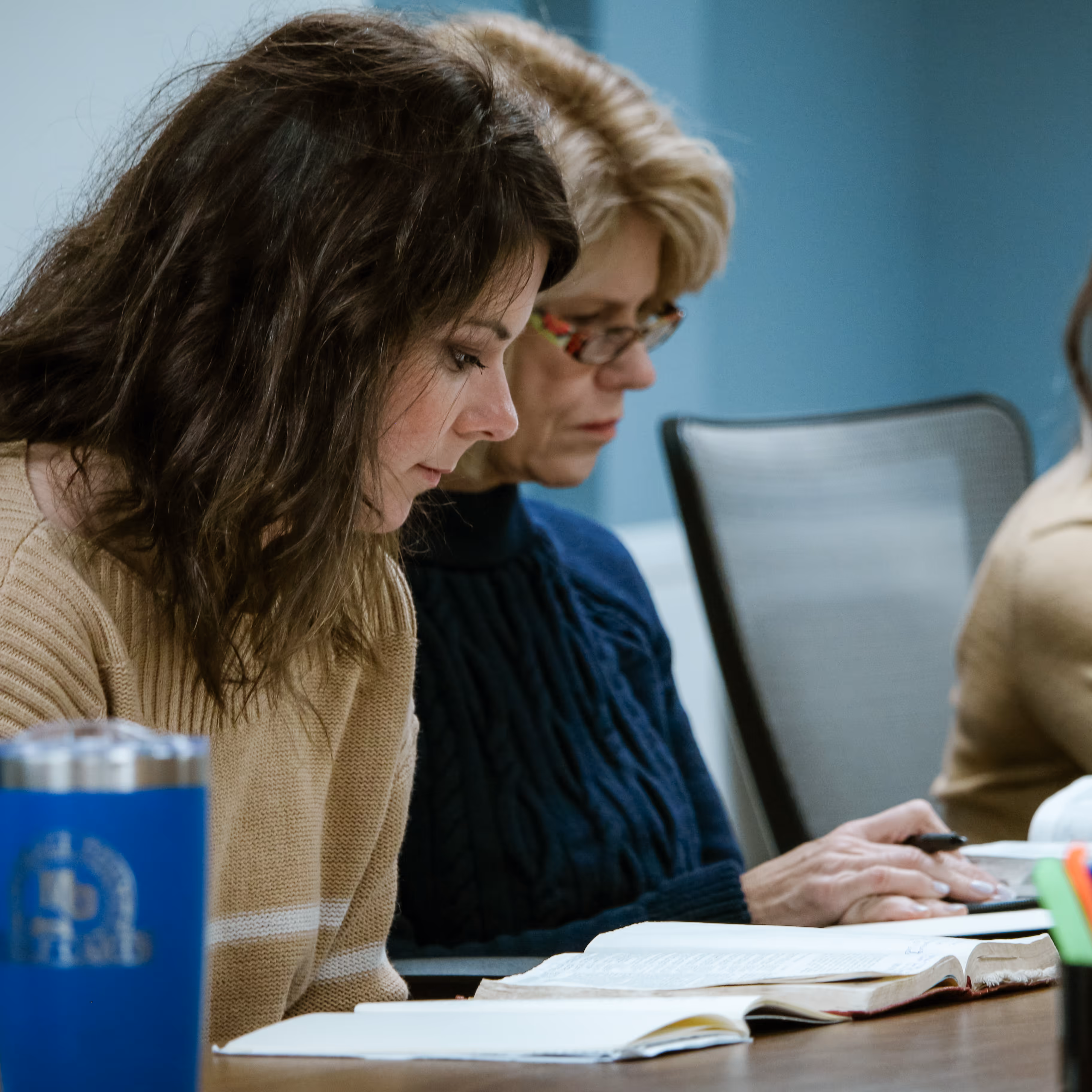 Two women seated at a table, reading open Bibles and taking notes.